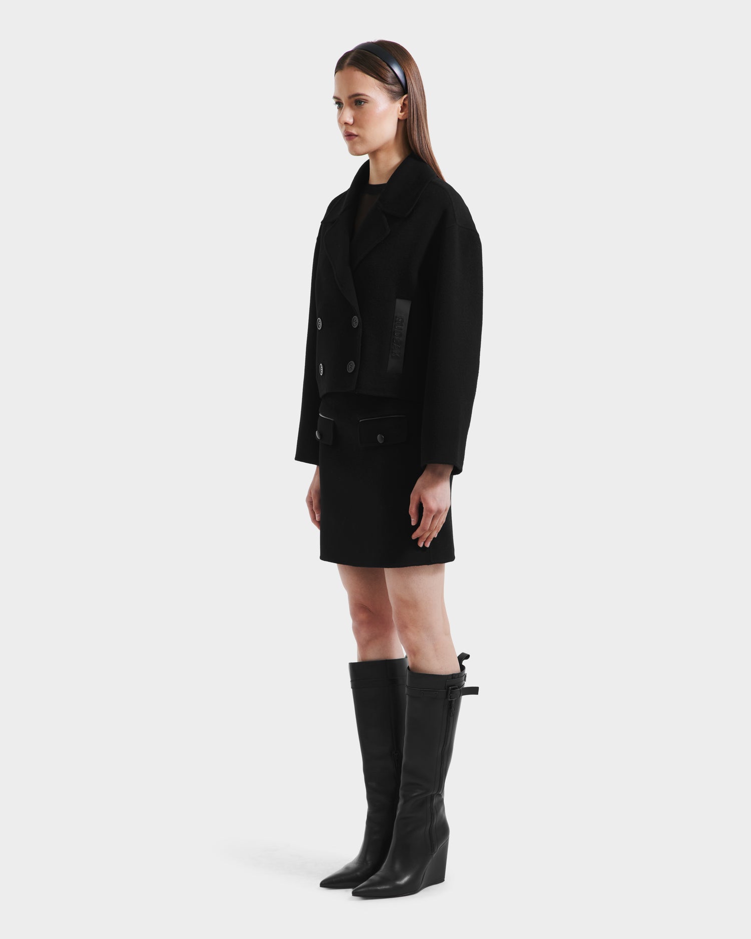 A woman in profile wears the Rudsak MARTINEZ - 8125580 Black vegan leather double-breasted jacket, paired with a black skirt and knee-high boots. Her straight brown hair is styled with a black headband against a plain light background.