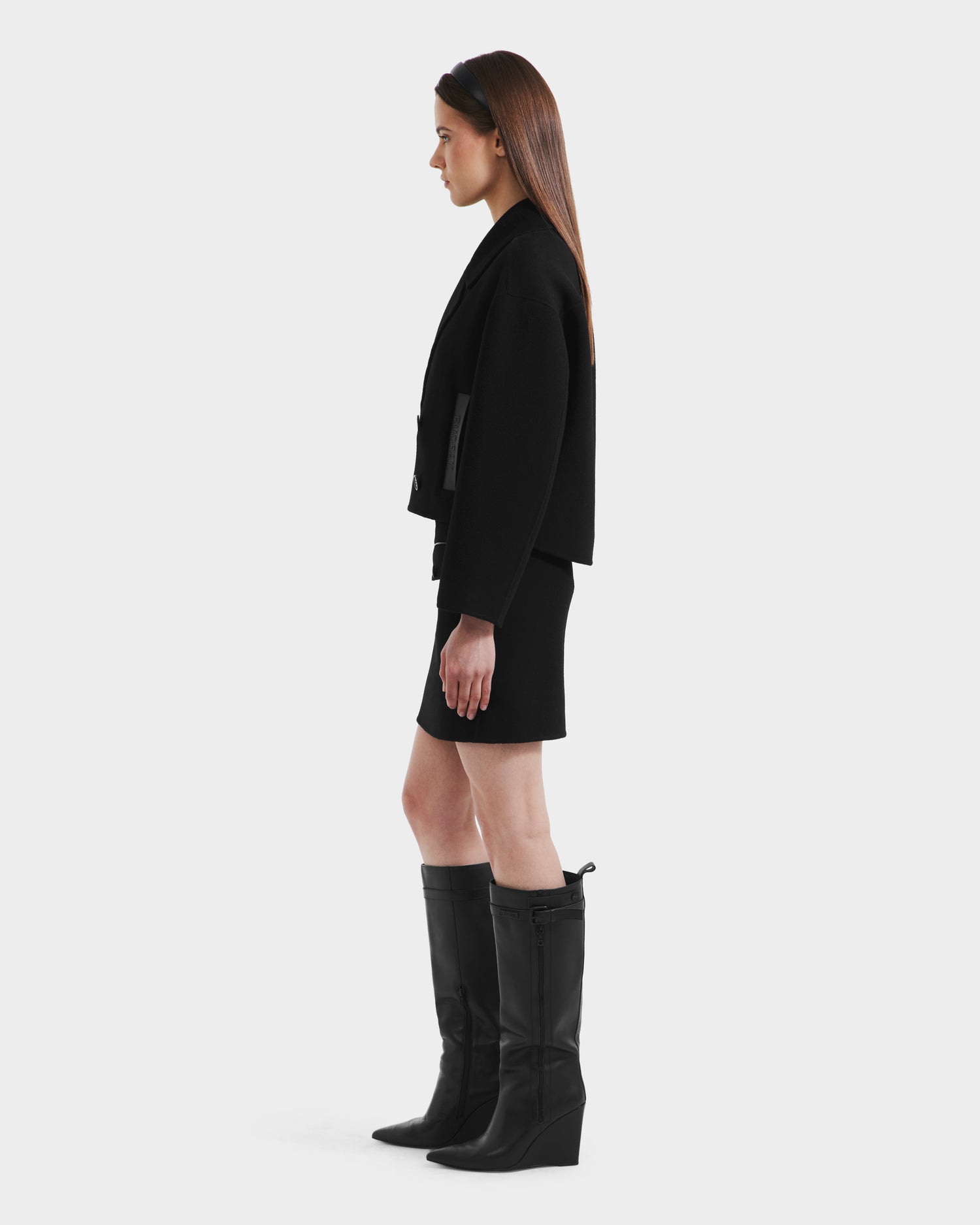 A woman with long straight hair stands in profile wearing the Rudsak MARTINEZ - 8125580 Black short coat, paired with a black skirt and knee-high boots, against a plain light background.