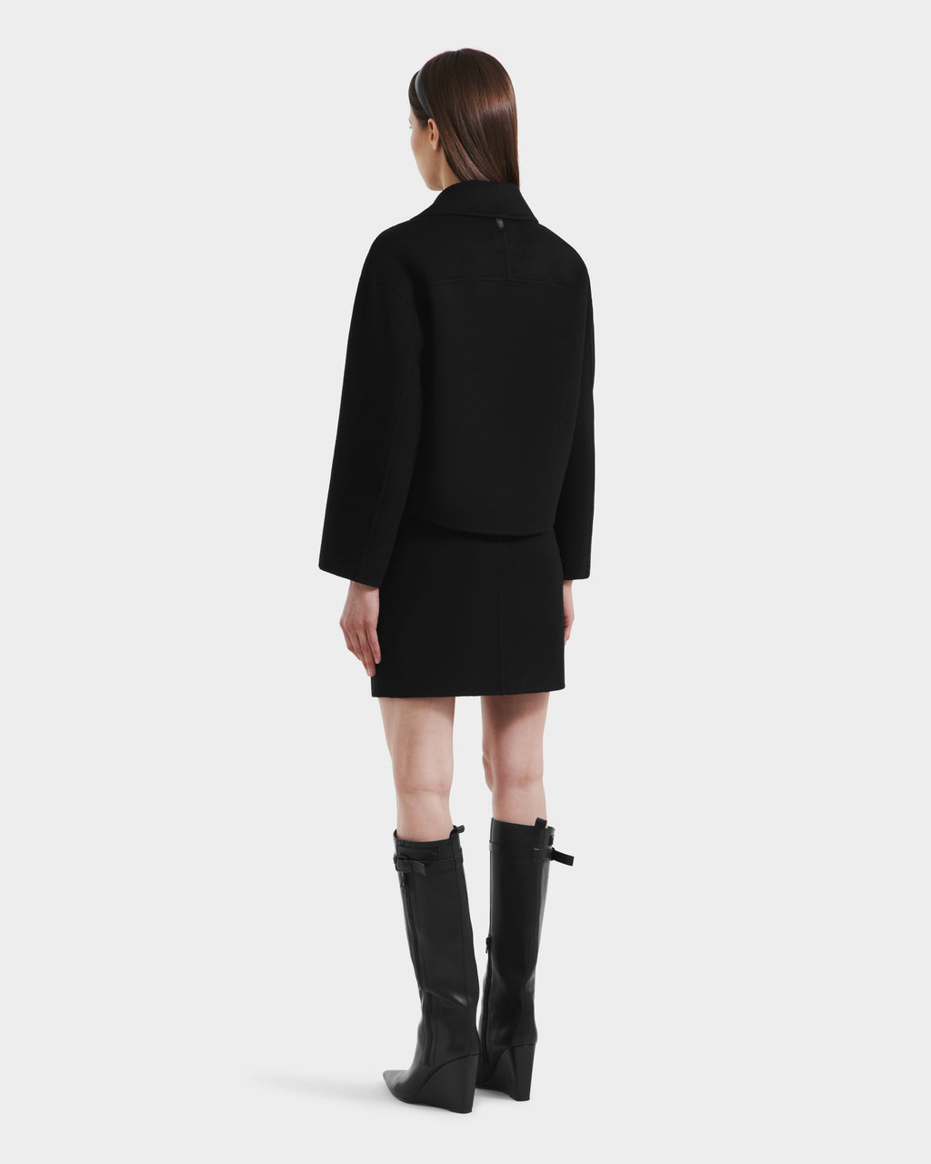 A woman with straight brown hair poses against a white background, wearing the Rudsak MARTINEZ - 8125580 Black vegan leather jacket, matching black skirt, and knee-high black boots with buckles.