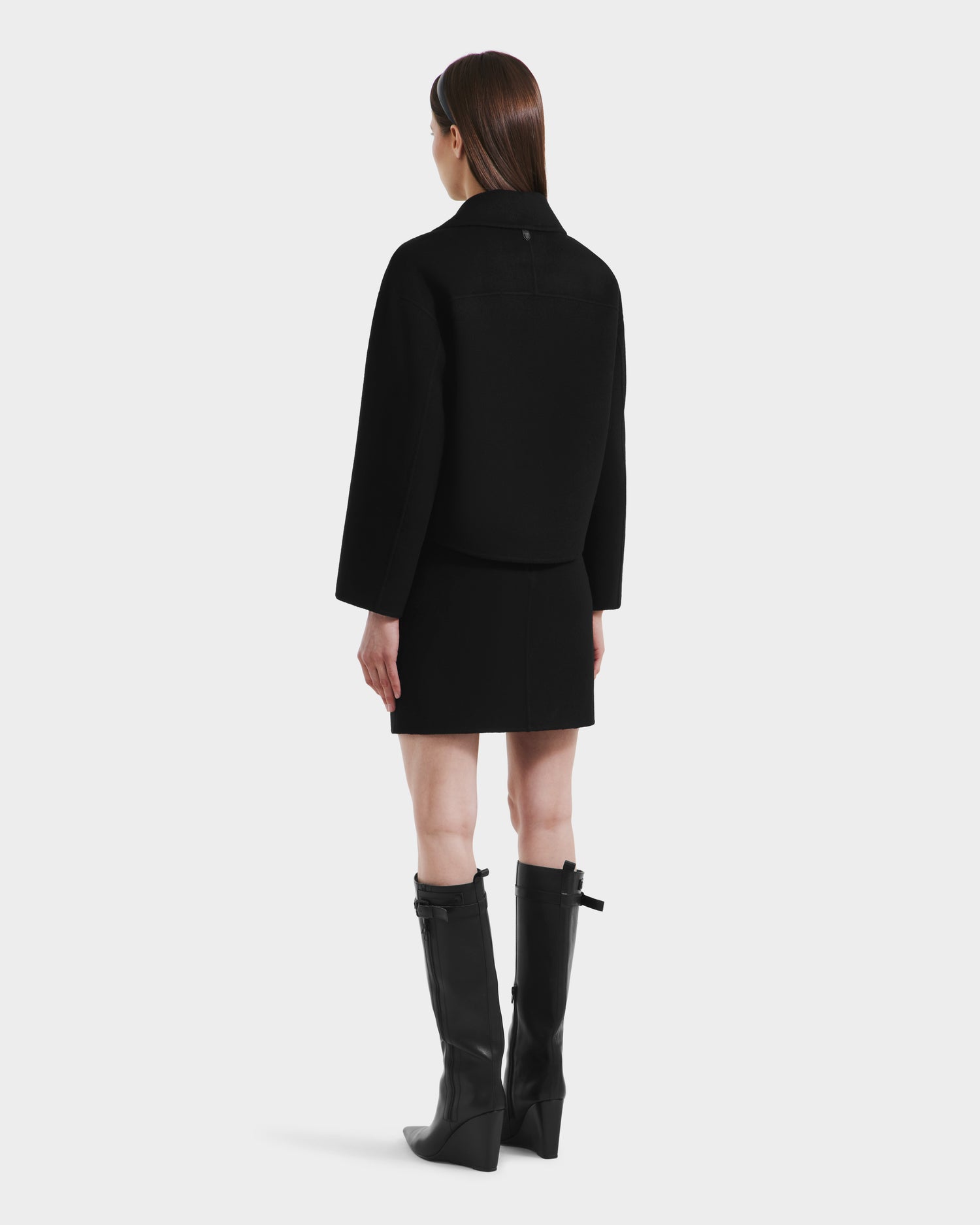 A woman with straight brown hair poses against a white background, wearing the Rudsak MARTINEZ - 8125580 Black vegan leather jacket, matching black skirt, and knee-high black boots with buckles.
