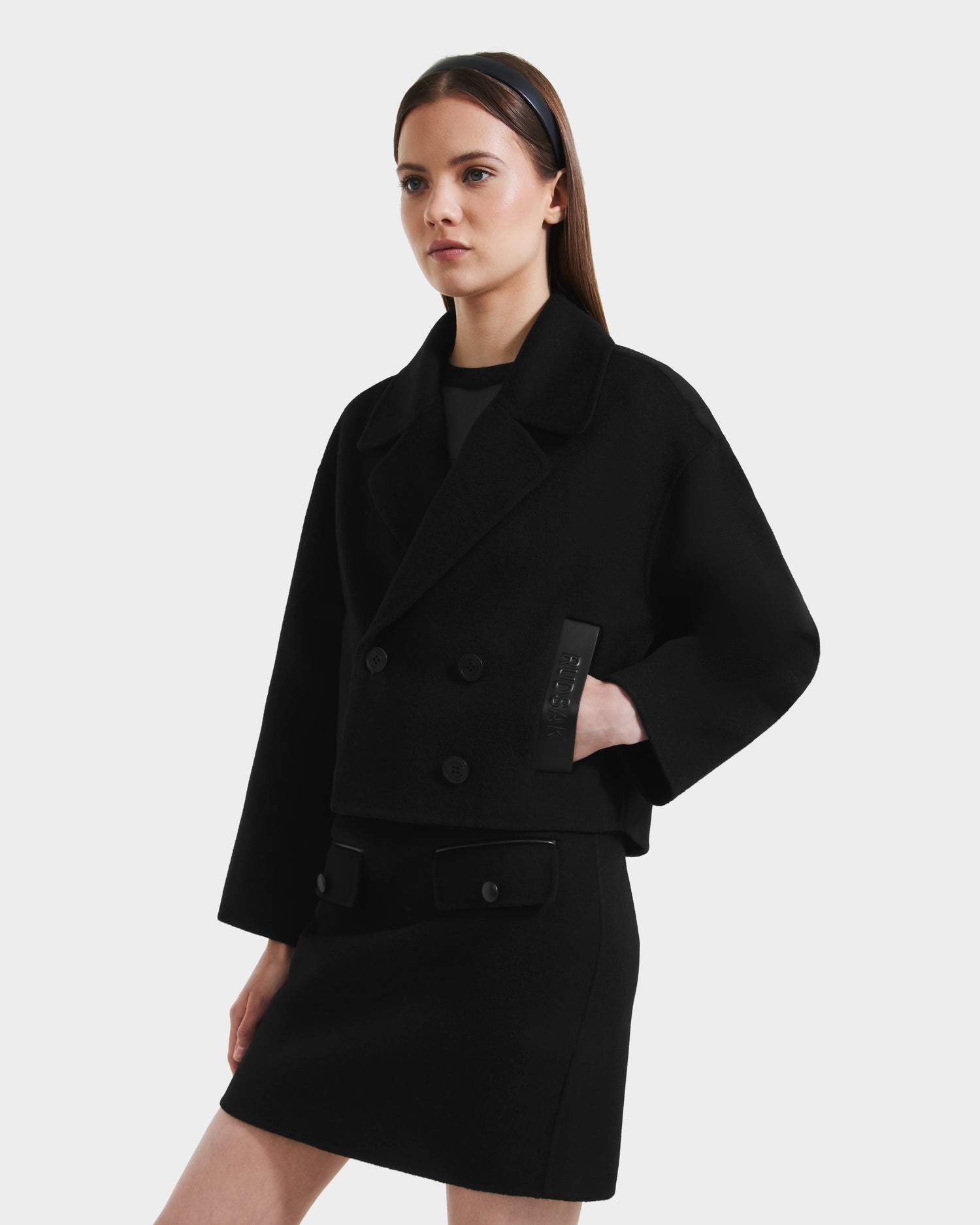 A woman stands against a plain light background wearing the Rudsak MARTINEZ - 8125580 Black double-breasted vegan leather cropped jacket with large lapels and a matching black skirt.