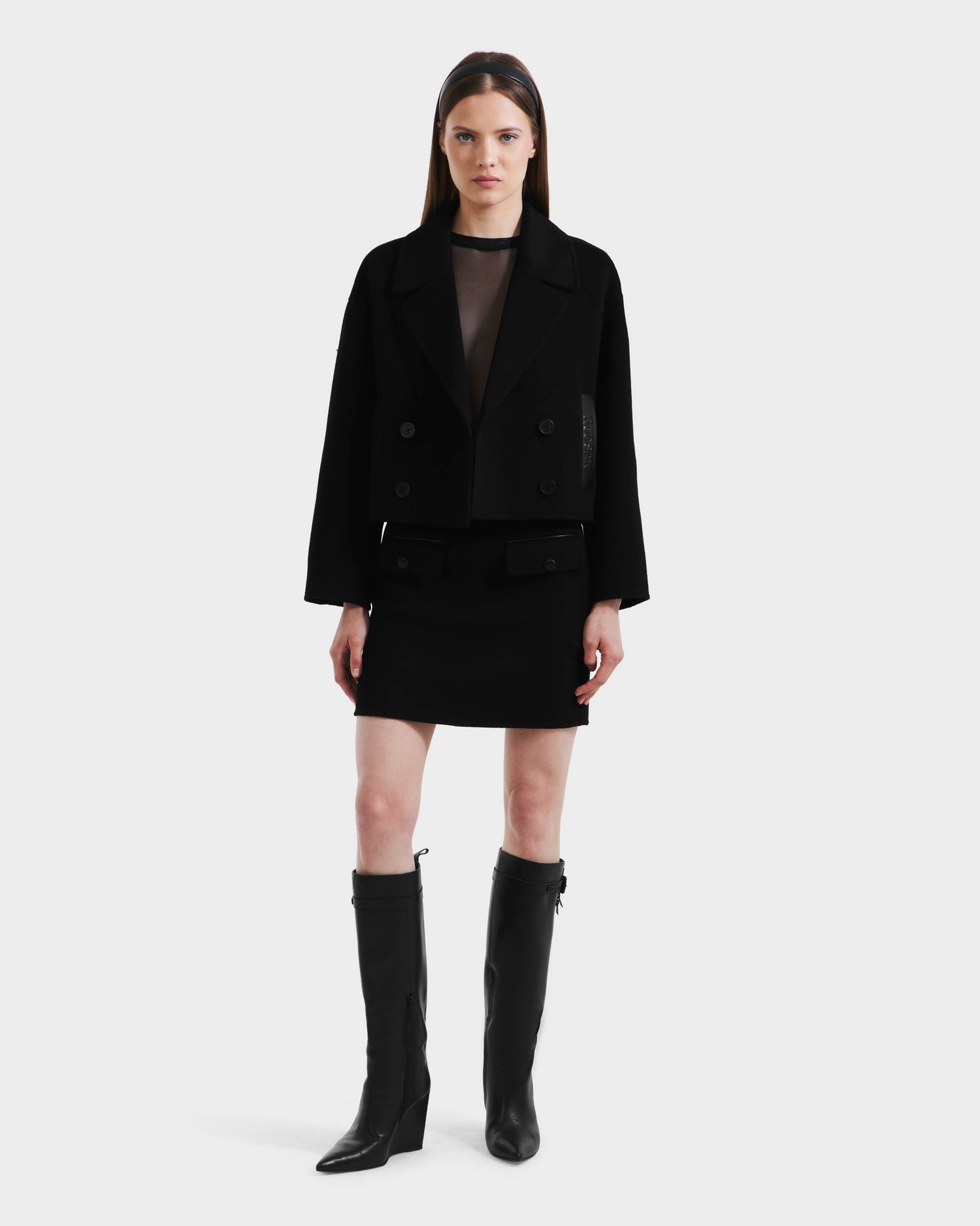 A woman faces forward against a white background, wearing the Rudsak MARTINEZ - 8125580 Black vegan leather cropped jacket, a sheer black top, black mini skirt, and knee-high black boots.