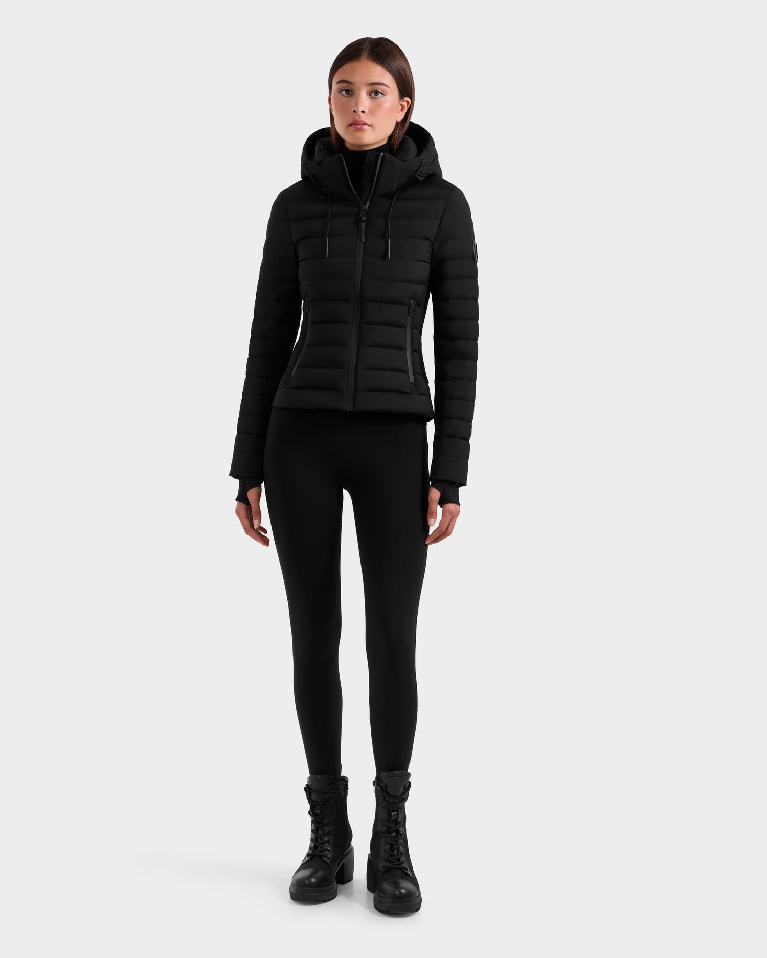 A woman faces forward wearing the Rudsak MARYA M - L125509 Black fitted down puffer jacket with RDS certified down, paired with black leggings and lace-up boots, set against a plain light gray background.