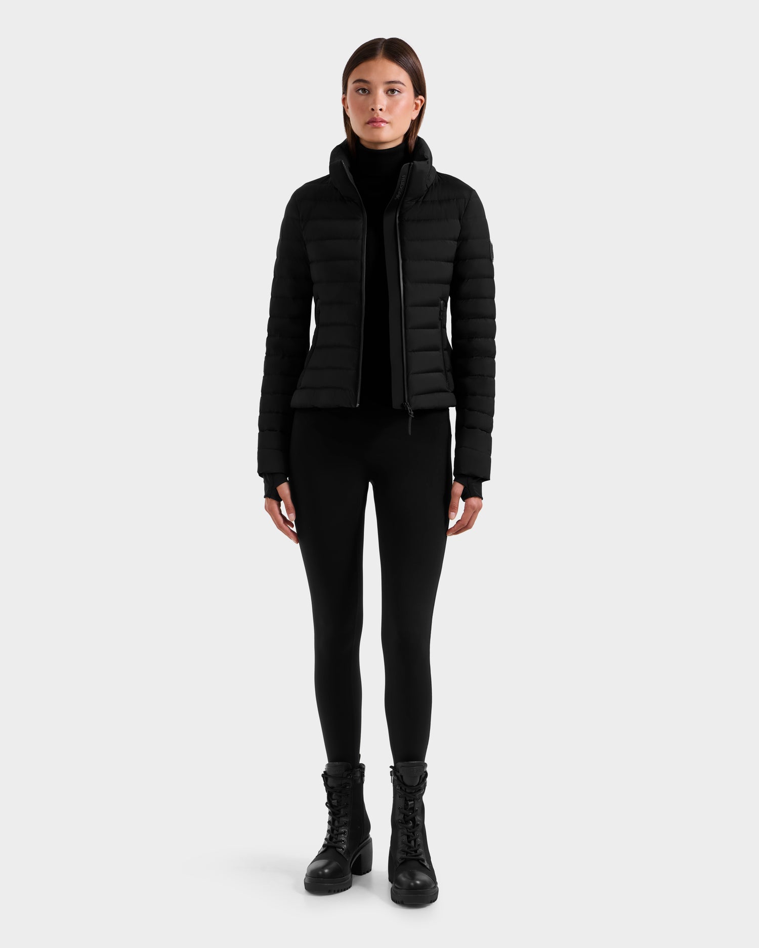 A woman faces forward wearing the Rudsak MARYA M - L125509 Black women’s down puffer jacket with RDS certified down, a black turtleneck, black leggings, and black lace-up boots against a plain white background.