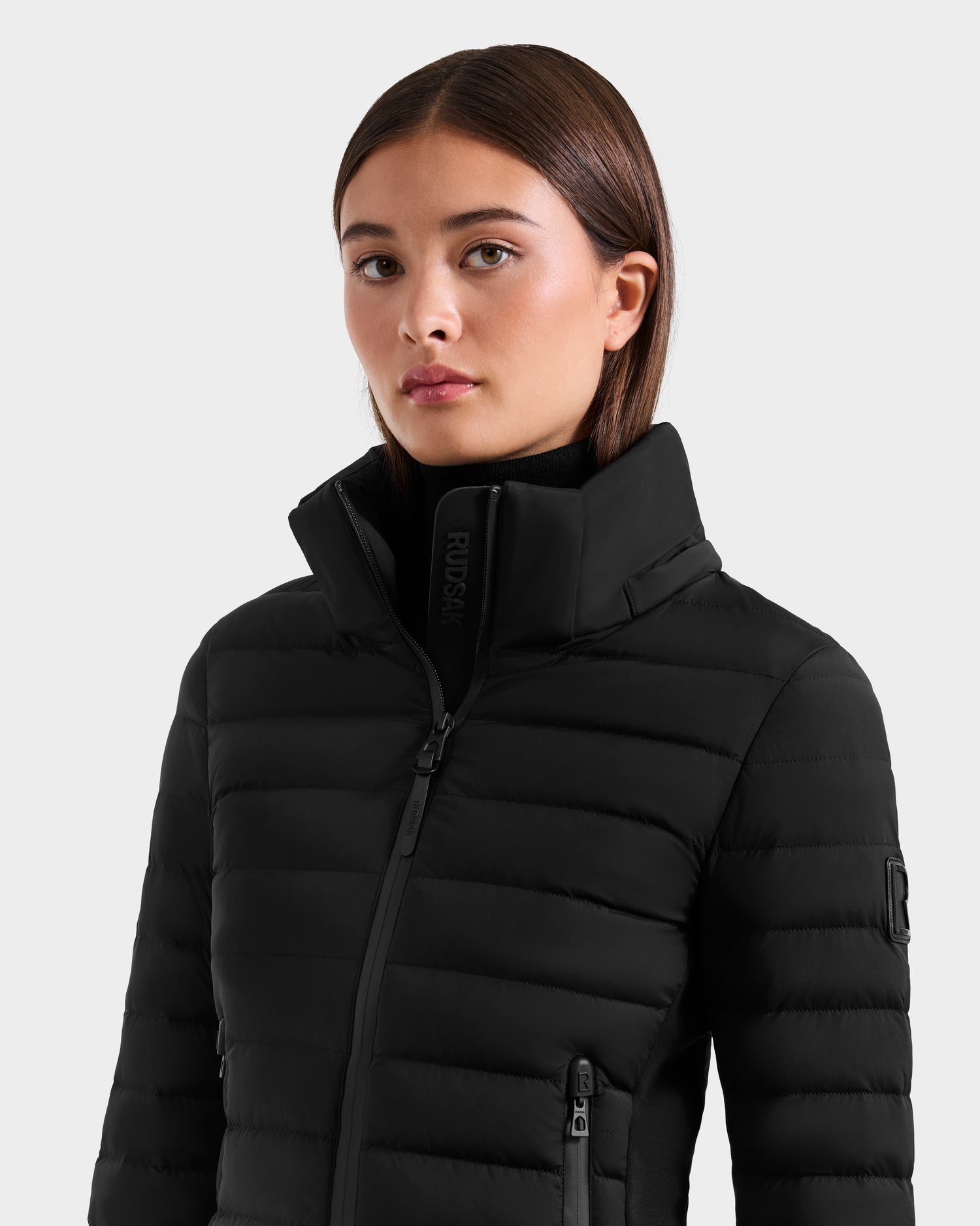 A young woman with straight brown hair wears the Rudsak MARYA M - L125509 Black women’s down puffer jacket with a high collar, looking slightly to the side with a neutral expression against a plain light gray background.