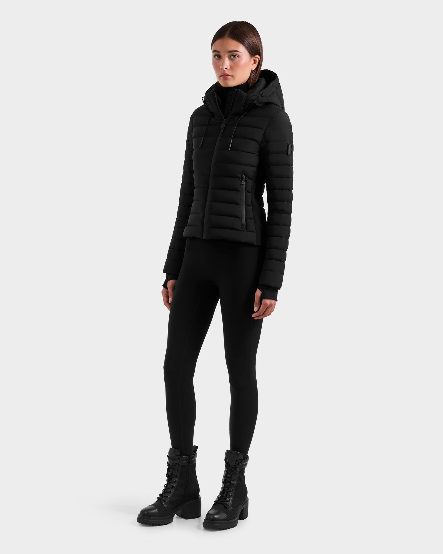 A woman stands against a plain background wearing the Rudsak MARYA M - L125509 Black fitted women’s down puffer jacket, paired with black leggings and lace-up boots, looking slightly to the side with relaxed arms.