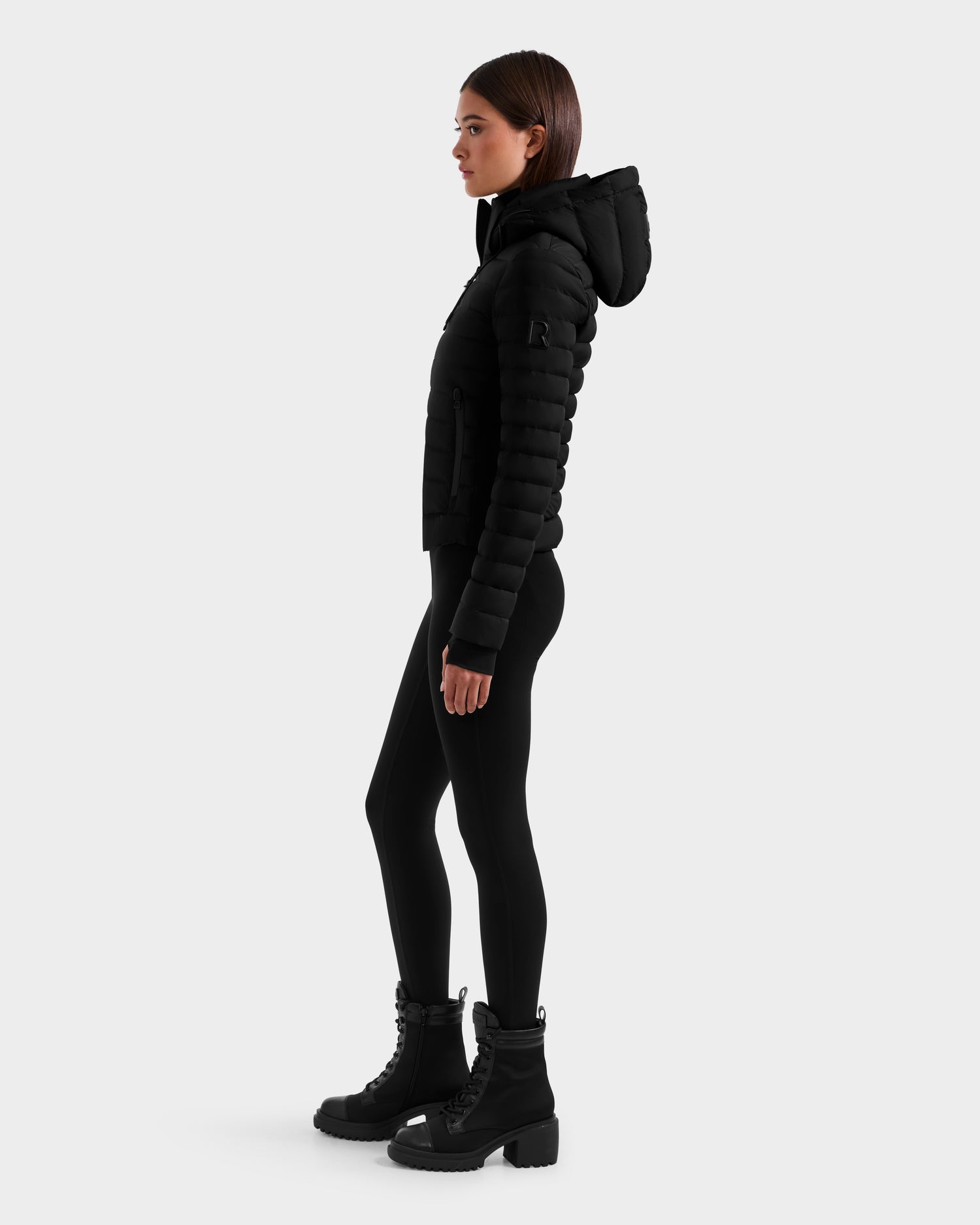 A woman stands in profile against a light gray background wearing the Rudsak MARYA M - L125509 Black down puffer jacket with a hood, paired with black leggings and wind- and water-repellent lace-up boots.
