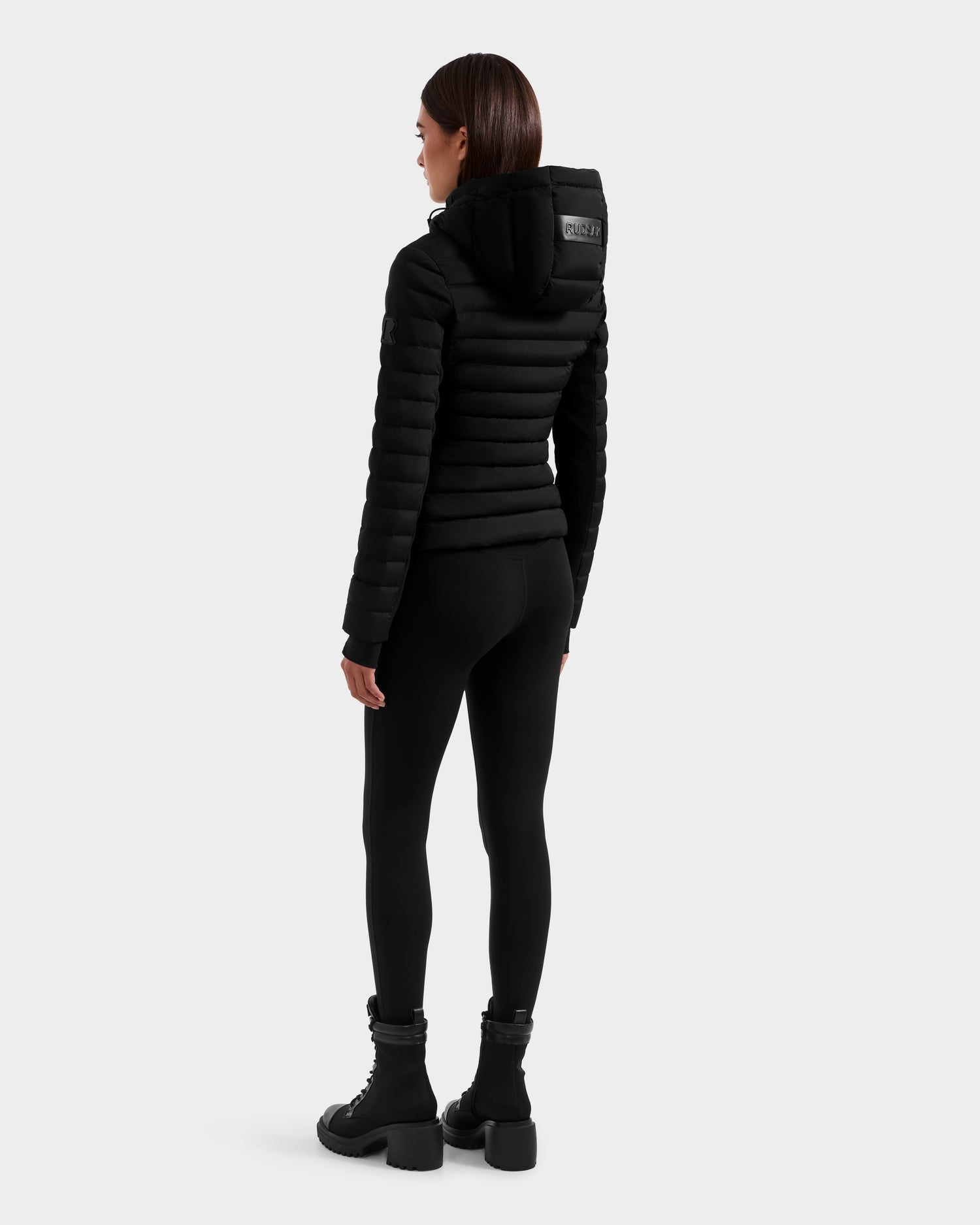A woman faces away, wearing the Rudsak MARYA M - L125509 Black fitted women’s down puffer jacket, paired with black leggings and ankle boots, set against a plain light gray background.