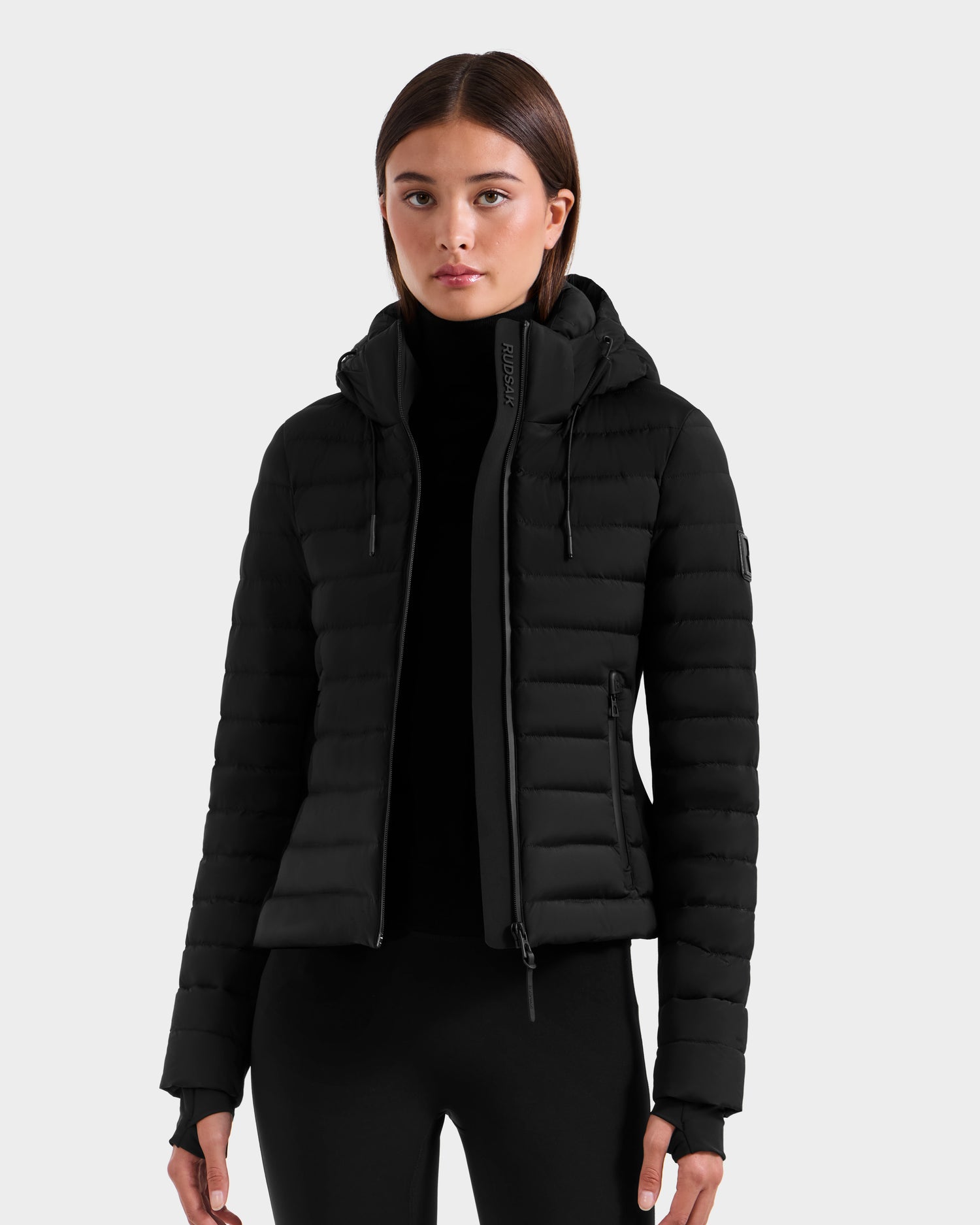 A young woman stands facing forward against a plain light gray background, wearing the Rudsak MARYA M - L125509 Black women's down puffer jacket with hood and zipper pockets, paired with black pants and a black turtleneck.