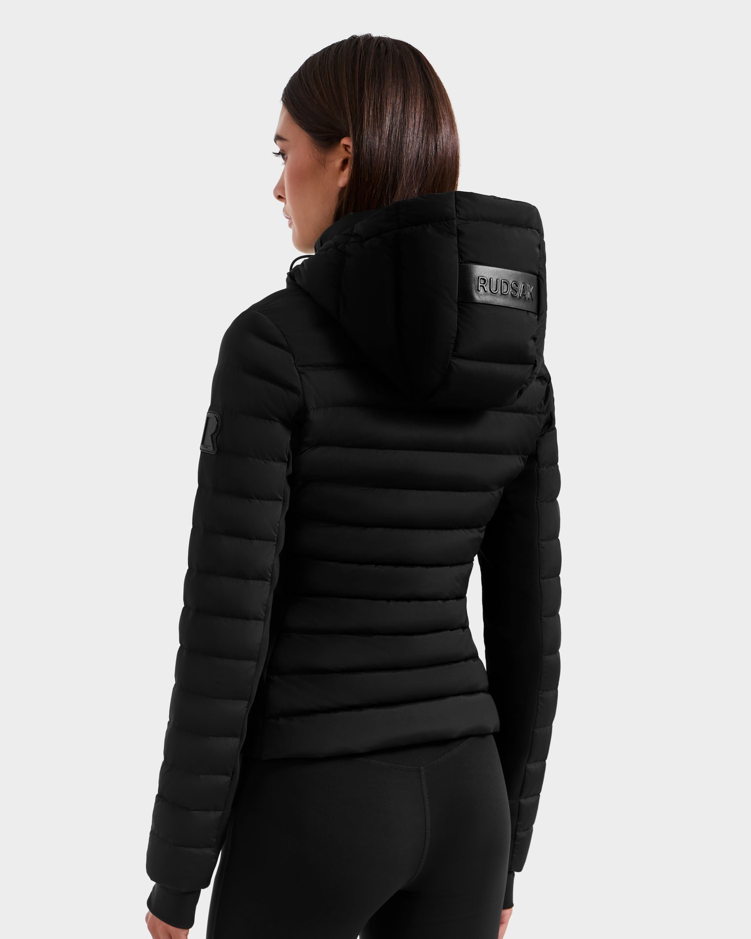 A woman with straight brown hair, seen from the back, wears the Rudsak MARYA M - L125509 Black fitted women’s down puffer jacket with hood, padded horizontal sections, wind and water-repellent finish, and a patch on the upper hood.