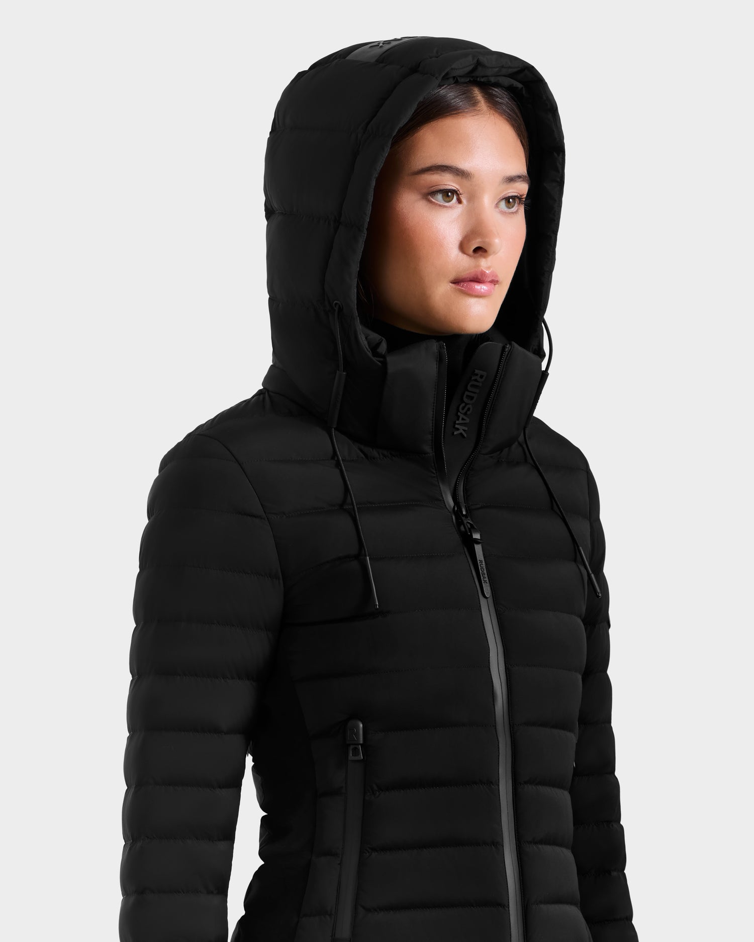 A woman wearing the Rudsak MARYA M - L125509 Black puffer jacket stands against a plain light grey background, looking slightly to the side with a neutral expression.