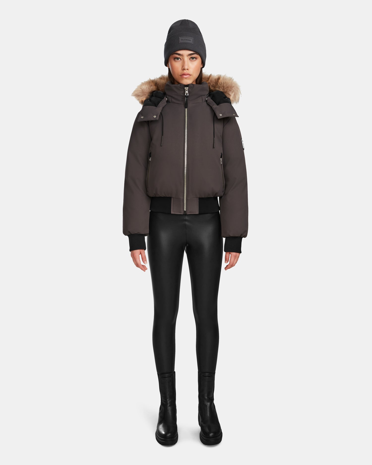 A woman faces forward against a white background, wearing the Rudsak MAUDE - L124522 Grey jacket—a dark brown puffer with premium down and fur-lined hood—paired with a gray beanie, black leggings, and black boots.