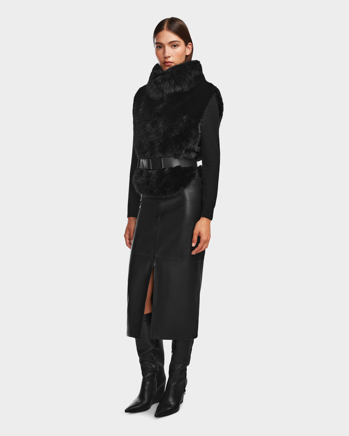 A woman poses against a plain background wearing the Rudsak MAULIN - 8524611 Black leather midi skirt with a front slit, styled with a chic pullover vest, black long-sleeve top, and black knee-high boots.