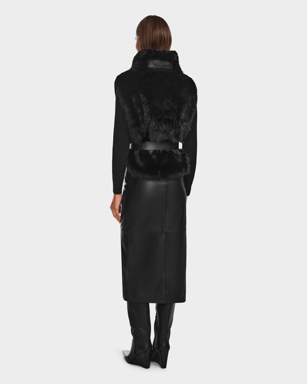 A woman with straight hair stands facing away against a light gray backdrop, wearing the Rudsak MAULIN - 8524611 Black faux fur vest over a black top, black leather skirt, and knee-high boots.