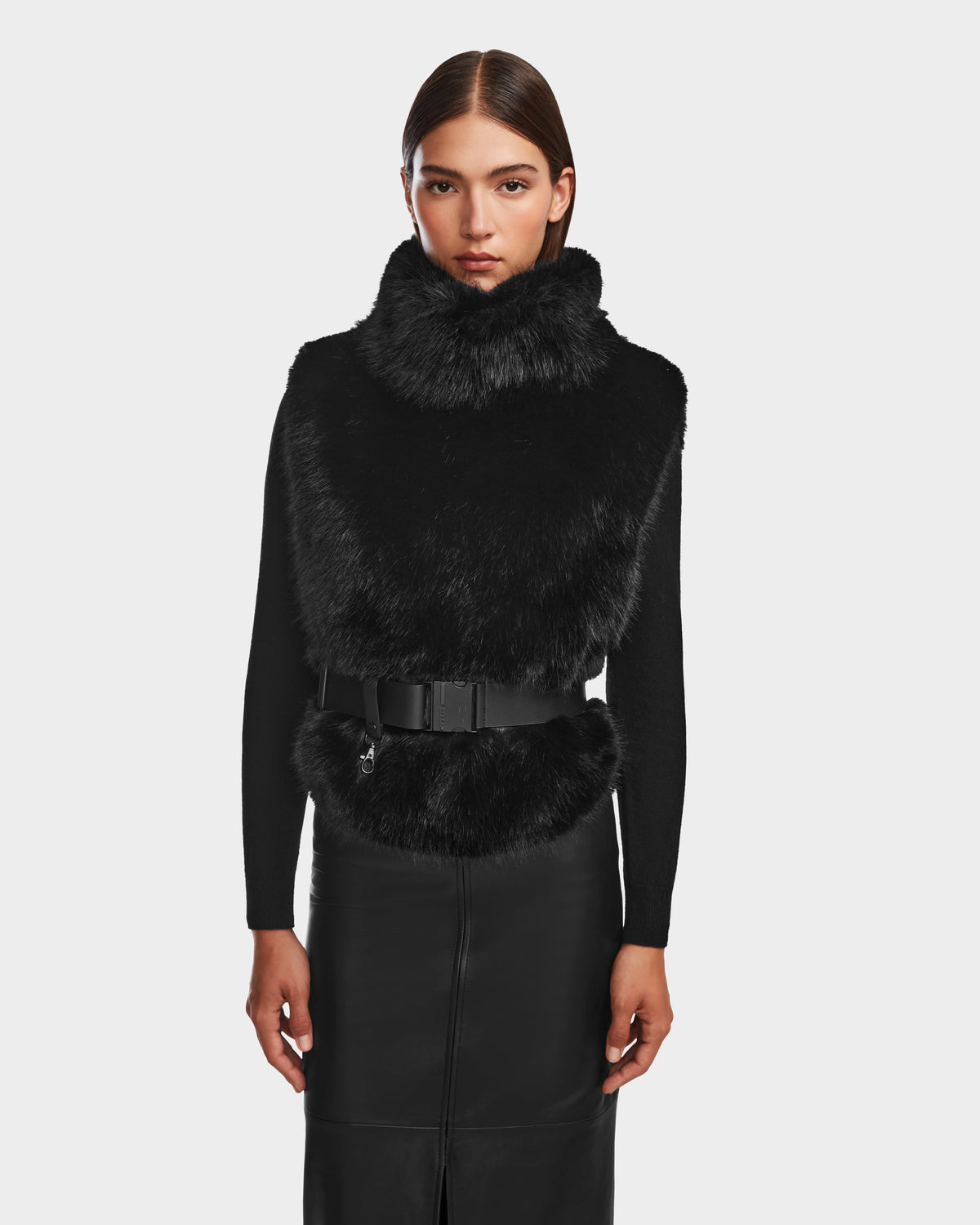 A woman with straight brown hair wears the Rudsak MAULIN - 8524611 Black faux fur vest over a black mock-neck pullover and a belted vegan leather skirt, standing against a plain light gray background.