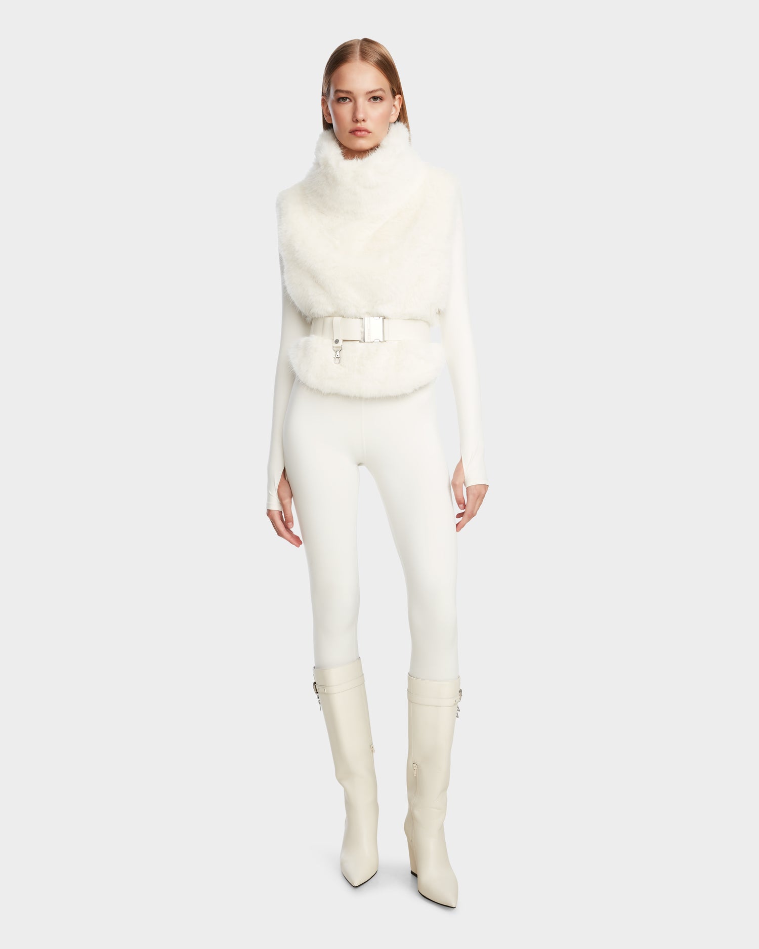 A woman faces forward in Rudsak’s MAULIN - 8524611 Iceland: a sleeveless faux fur belted vest, fitted long-sleeve top and pants, and knee-high vegan leather pointed-toe boots with block heels. Background: plain light gray.