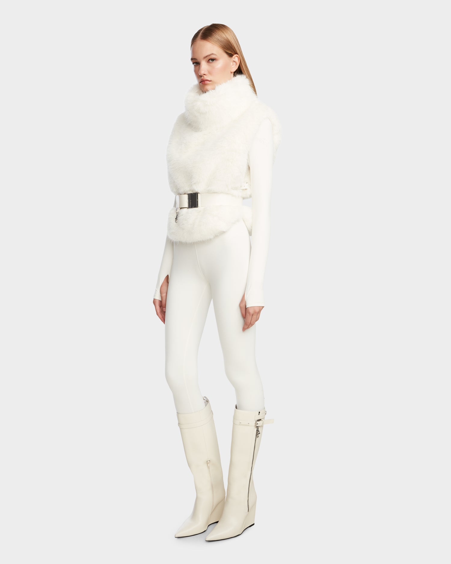 A person models the Rudsak MAULIN - 8524611 Iceland faux fur vest with a high neck, paired with white fitted pants, a wide silver belt, and tall white heeled boots against a plain light gray background.