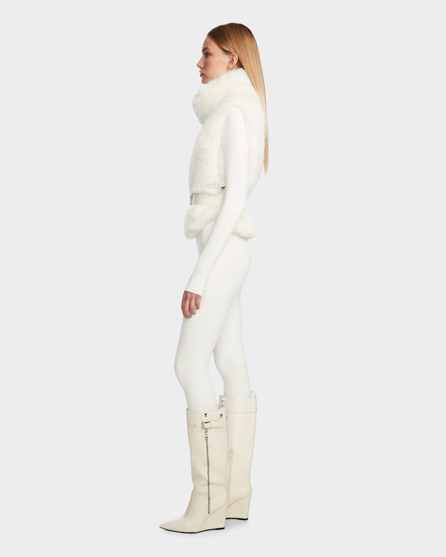 A woman in profile wears the Rudsak MAULIN - 8524611 Iceland chic faux fur vest over a fitted white bodysuit and off-white knee-high vegan leather boots with zippers, posed before a plain light gray background.