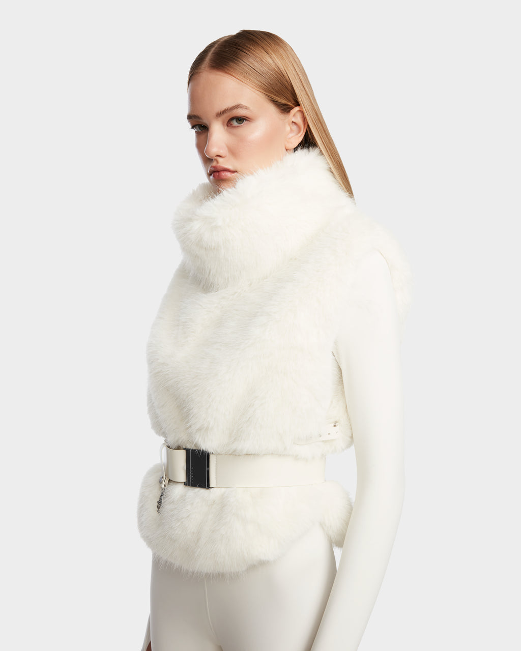 A woman with straight, light brown hair models the Rudsak MAULIN - 8524611 Iceland sleeveless white faux fur vest with a high collar and wide belt over a fitted vegan leather outfit against a plain light gray background.