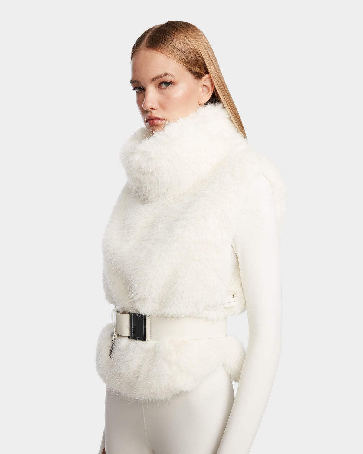 A woman with straight, light brown hair models the Rudsak MAULIN - 8524611 Iceland sleeveless white faux fur vest with a high collar and wide belt over a fitted vegan leather outfit against a plain light gray background.