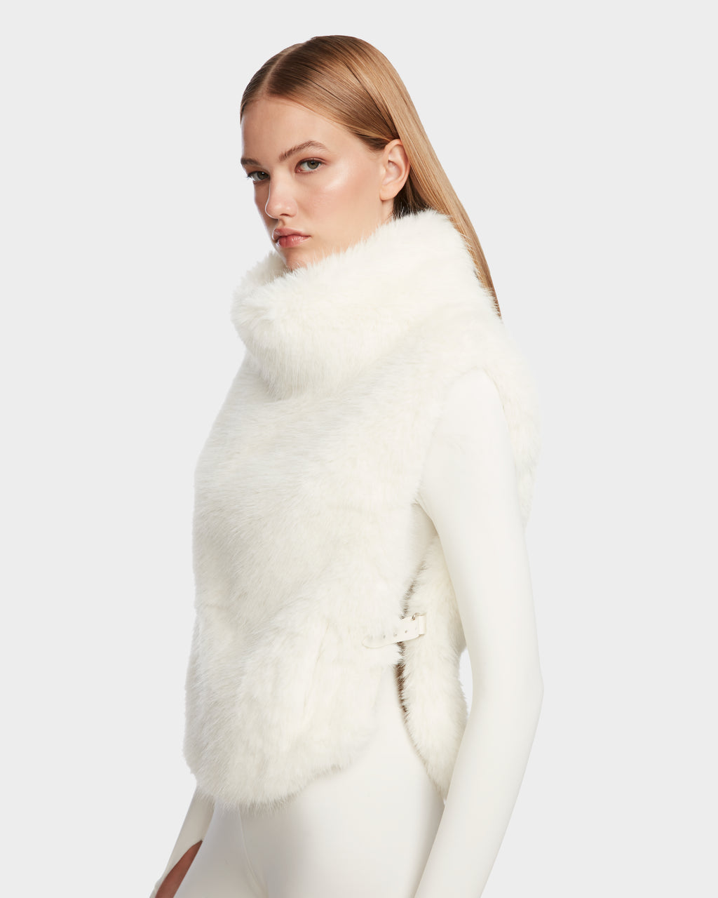 A woman with straight, light brown hair wears the Rudsak MAULIN - 8524611 Iceland faux fur vest with a high collar and side buckle over a fitted white outfit, posing stylishly against a light gray background.