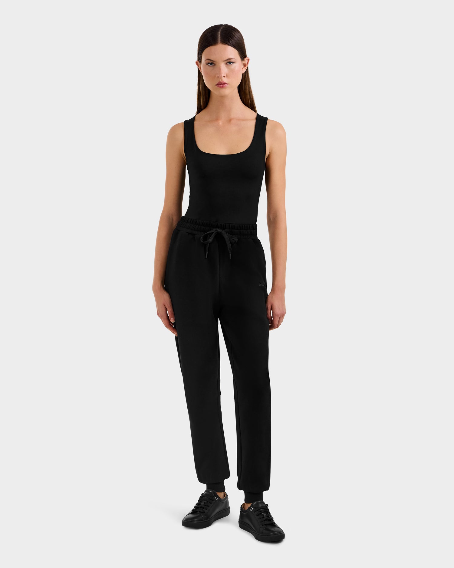 A woman with straight brown hair stands against a plain background wearing the Rudsak MAXTON - U525007 Black W sleeveless tank top and mid-weight fleece cotton-blend jogger pants with a drawstring, paired with black sneakers.