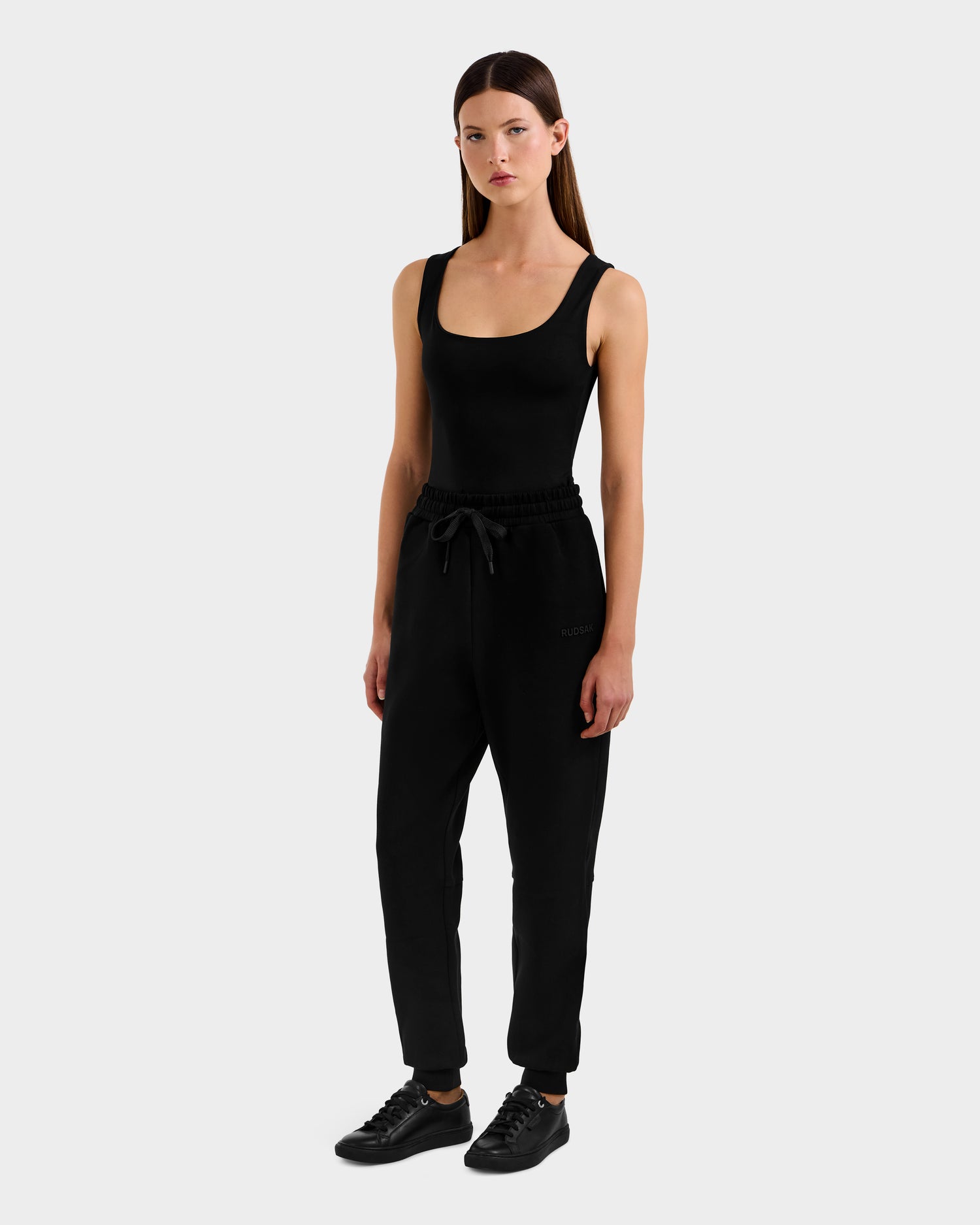 A woman faces forward, wearing the Rudsak MAXTON - U525007 Black W tank top with cotton-blend jogger pants and black sneakers, standing against a plain light gray background.
