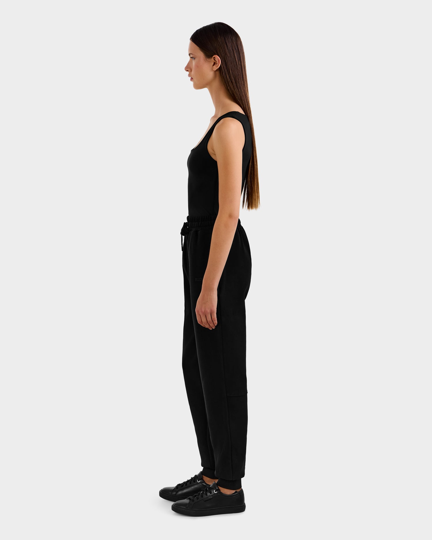 A woman with straight brown hair stands in profile, wearing the Rudsak MAXTON - U525007 Black W jogger pants and a sleeveless black top, against a plain light gray background.