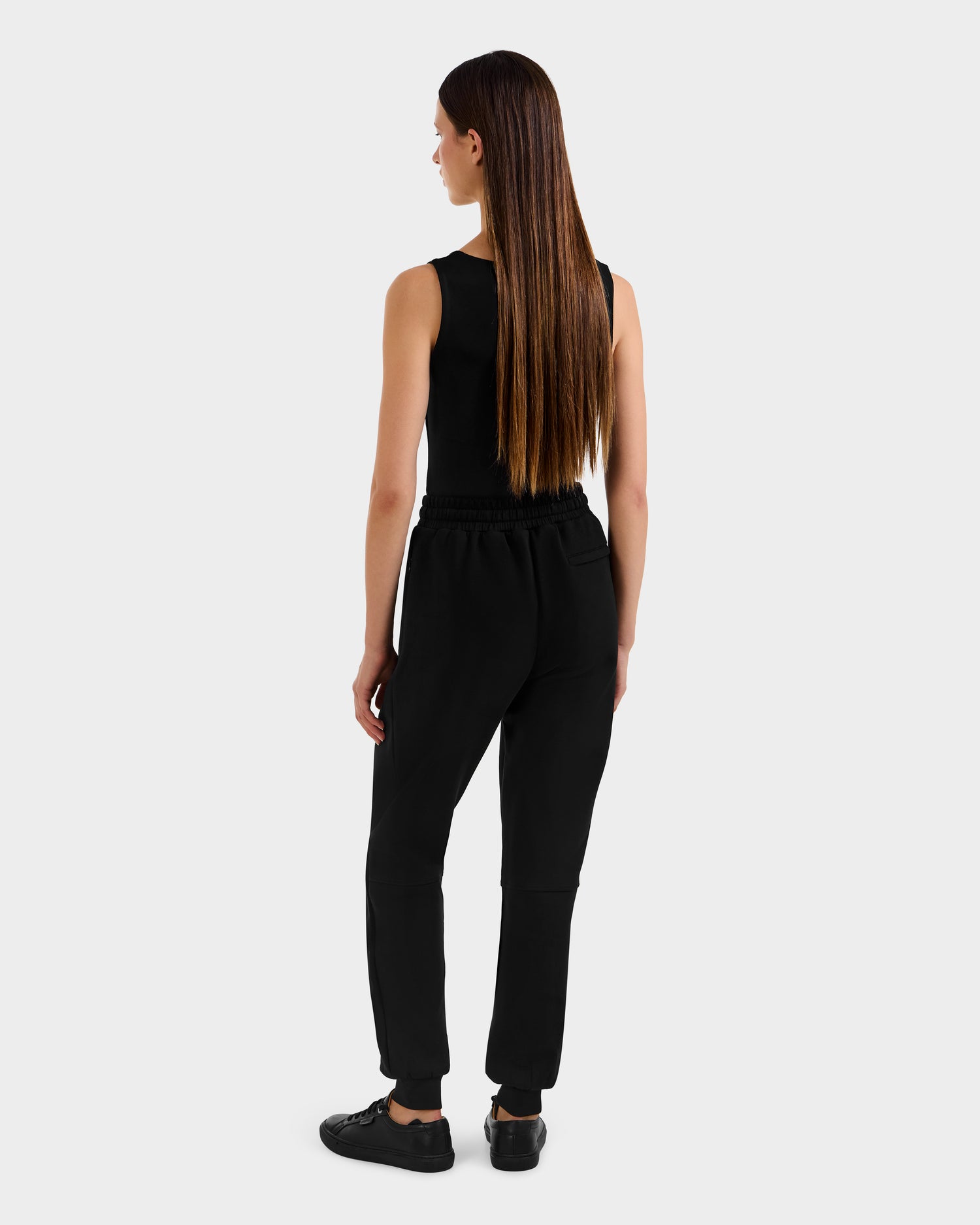 A woman with long straight brown hair, wearing Rudsak’s MAXTON - U525007 Black W jogger pants and a black sleeveless top, stands facing away against a plain light gray background.