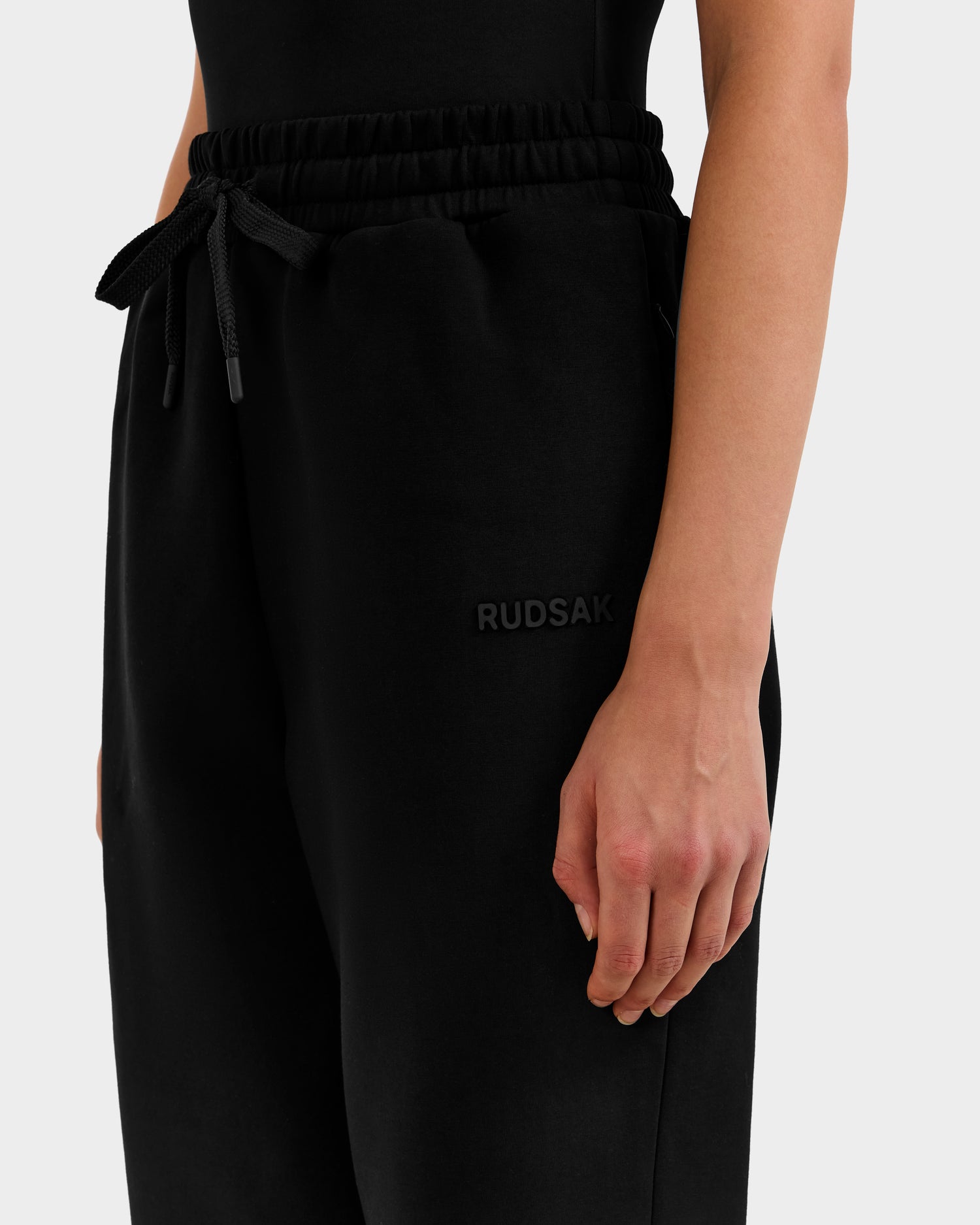 A person wears Rudsak's MAXTON - U525007 Black W jogger sweatpants, featuring a cotton-blend fabric, drawstring waist, and RUDSAK logo on the hip. The shot highlights the lower torso and right arm against a plain light background.