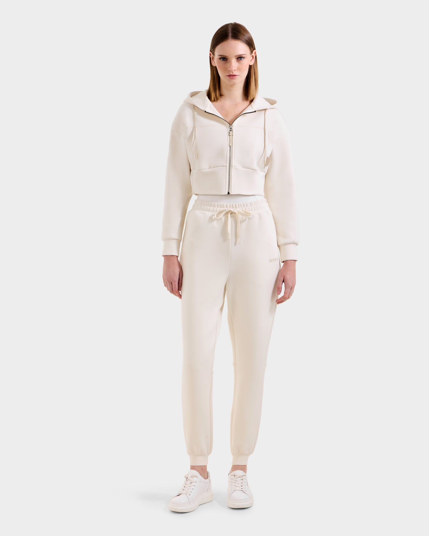 A woman stands facing forward, wearing the Rudsak MAXTON - U525007 Vanilla W zip-up hoodie and matching jogger pants, styled with white sneakers against a plain light background.
