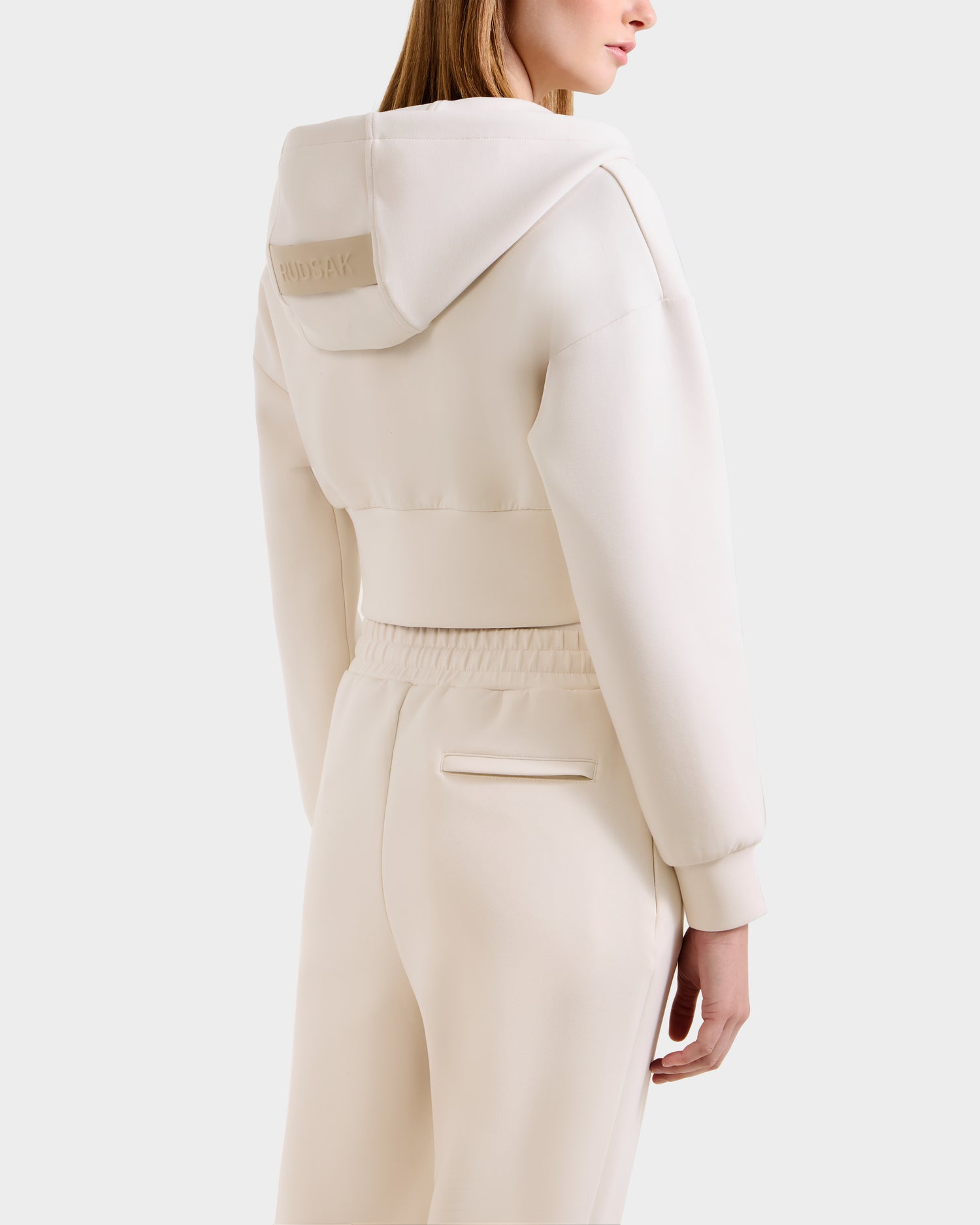 A woman wears the Rudsak MAXTON - U525007 Vanilla W cream hoodie and matching joggers, viewed from behind at an angle, displaying effortless everyday comfort against a plain white background.