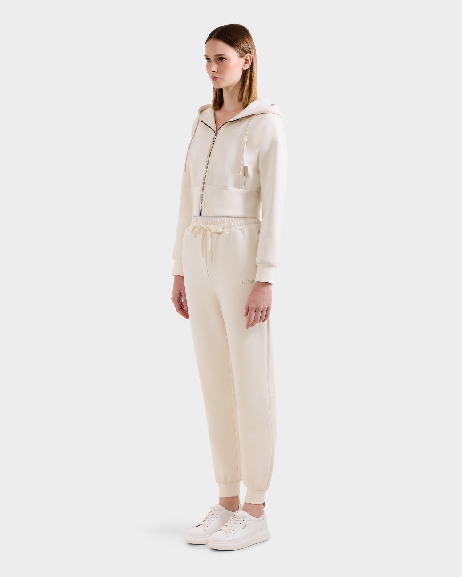 A woman wears the Rudsak MAXTON - U525007 Vanilla W off-white zip-up hoodie with matching jogger pants and white sneakers, facing slightly to the side against a plain light background, showcasing everyday comfort.