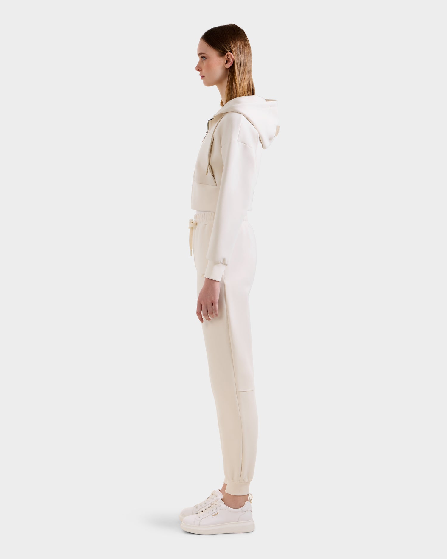 A woman stands in profile wearing the Rudsak MAXTON - U525007 Vanilla W jogger and hoodie set with white sneakers, showcasing everyday comfort against a plain white background.