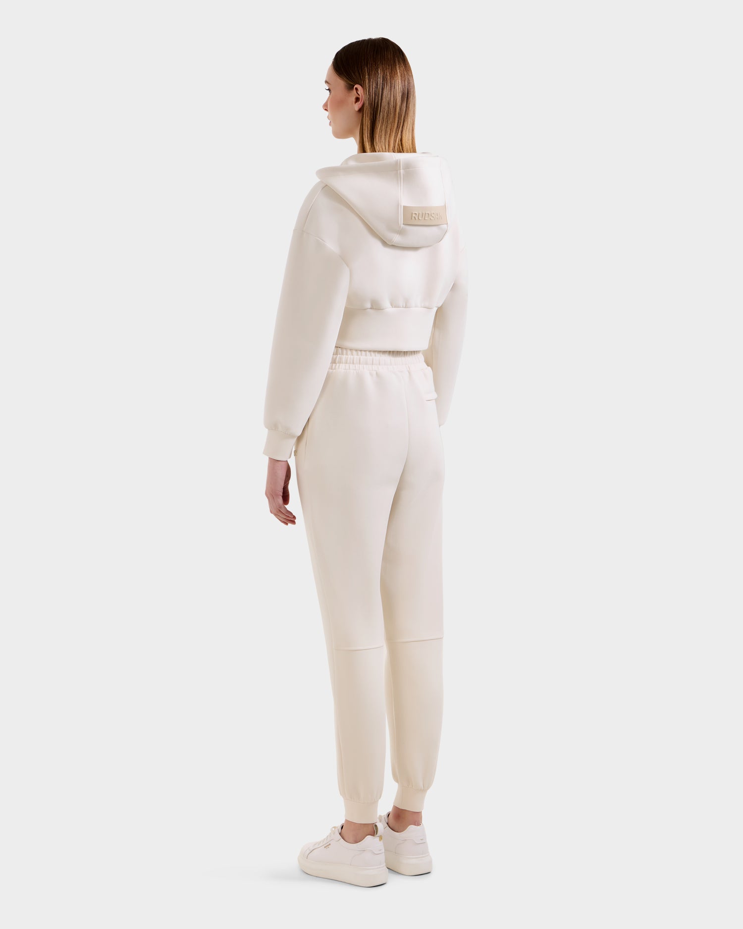 A person faces away, wearing Rudsak's MAXTON - U525007 Vanilla W off-white cropped hoodie and cotton-blend jogger sweatpants with white sneakers—an effortless, comfortable look against a plain light backdrop.