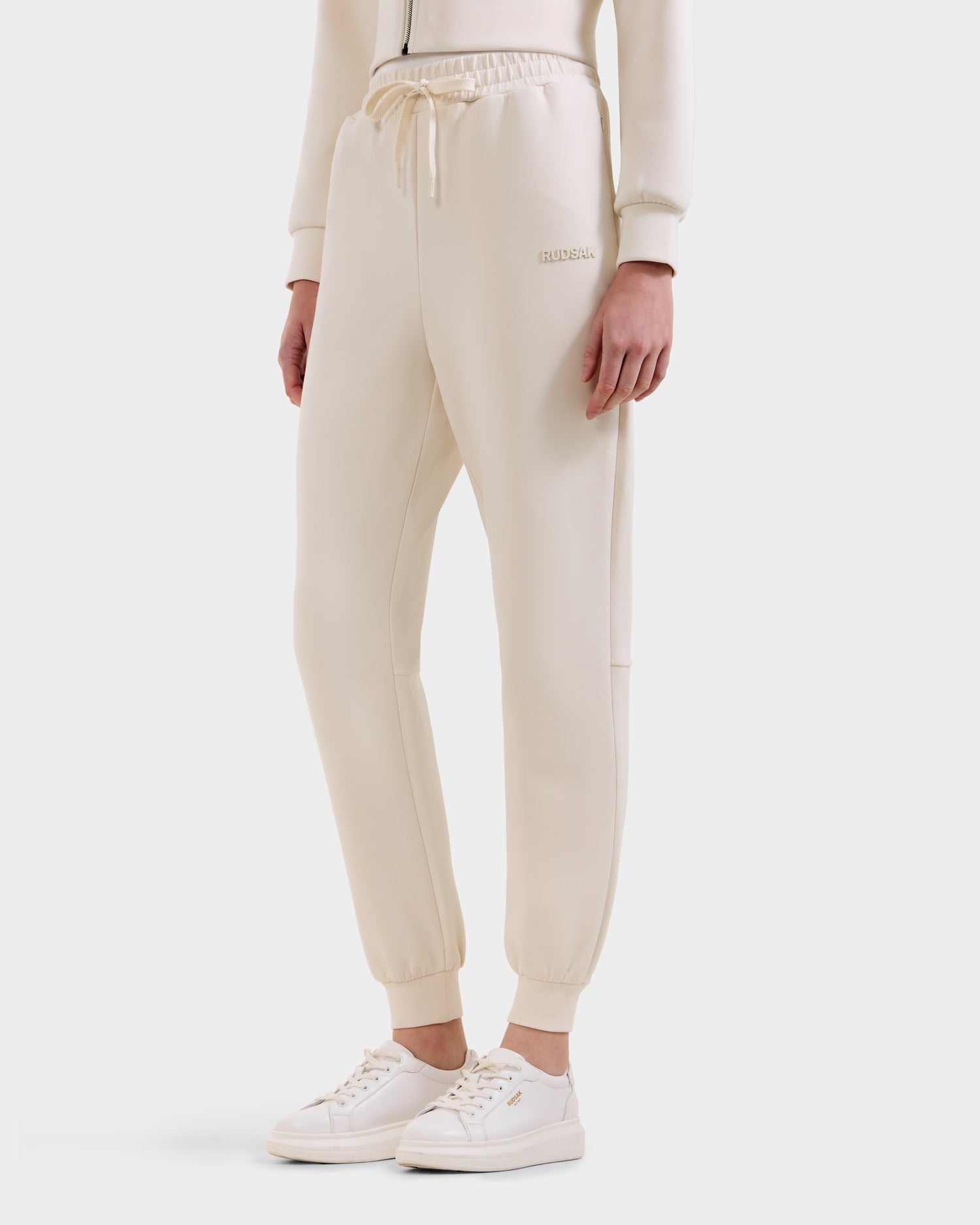 A person wears the Rudsak MAXTON - U525007 Vanilla W unisex cotton-blend jogger pants with an elastic waistband and drawstring, styled for everyday comfort, paired with matching sneakers and a hoodie against a plain white background.