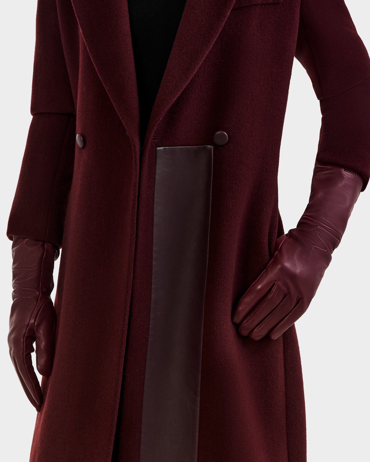 A woman wearing the Rudsak MEGAN - 8425541 Bordeaux long coat with dark buttons and matching bordeaux touchscreen leather gloves, shown from shoulders to knees against a plain background.