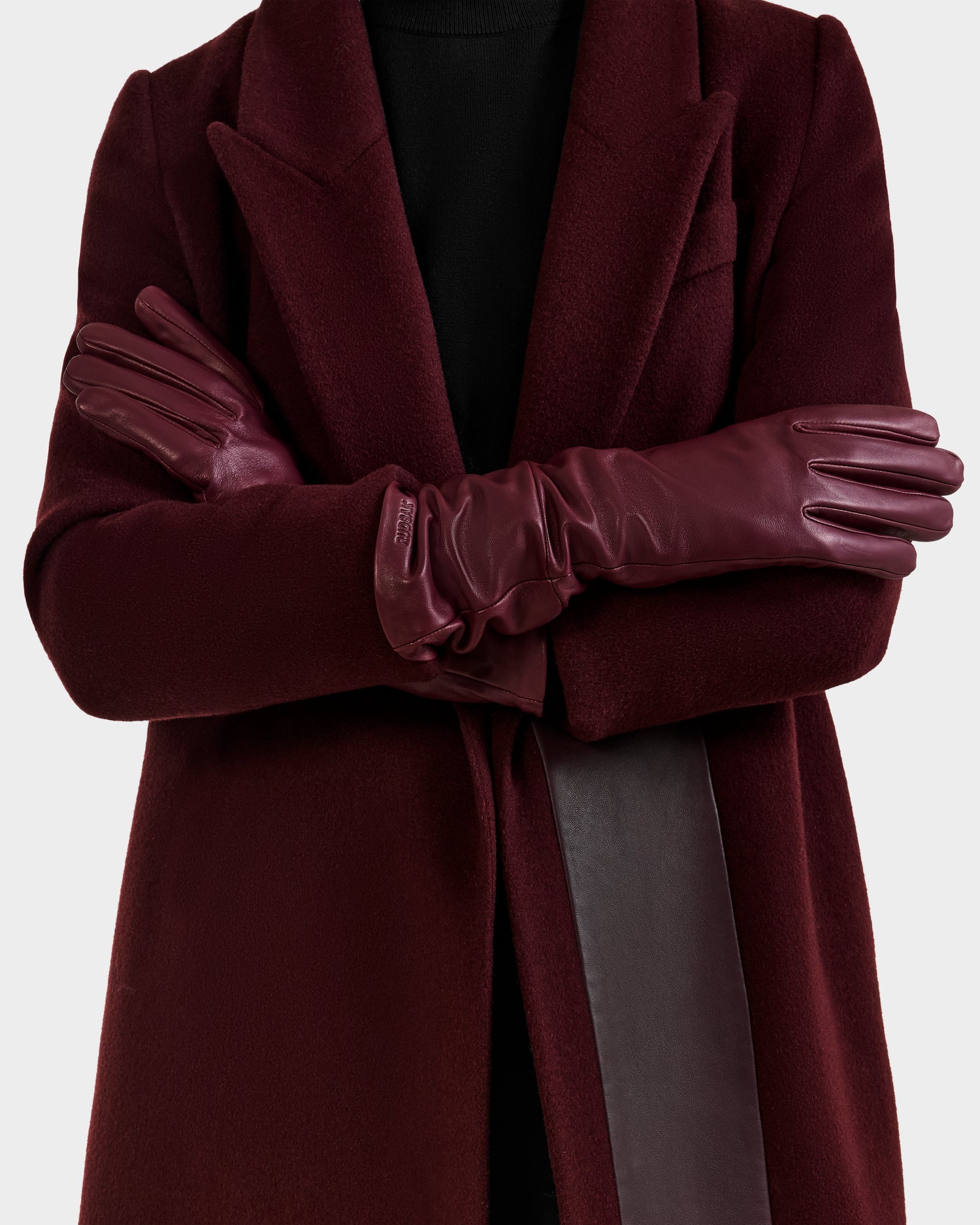 A person models the Rudsak MEGAN - 8425541 Bordeaux coat and touchscreen-compatible burgundy leather gloves, arms crossed, against a plain light background. Their face is not shown.