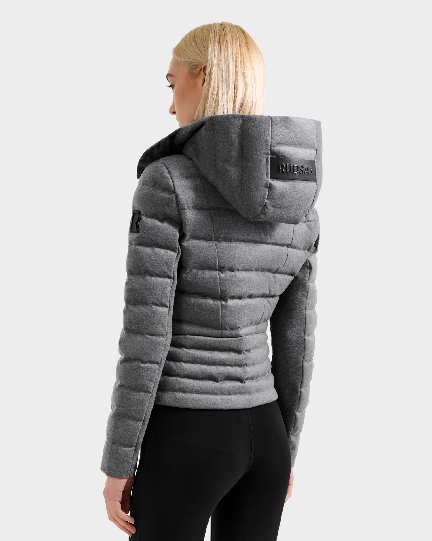 Seen from the back, a woman with straight blonde hair wears the MELISSA W - 8125564 Heather Charcoal fitted down jacket by Rudsak, featuring a hood with RUDSAK branding, paired with black leggings against a light background.