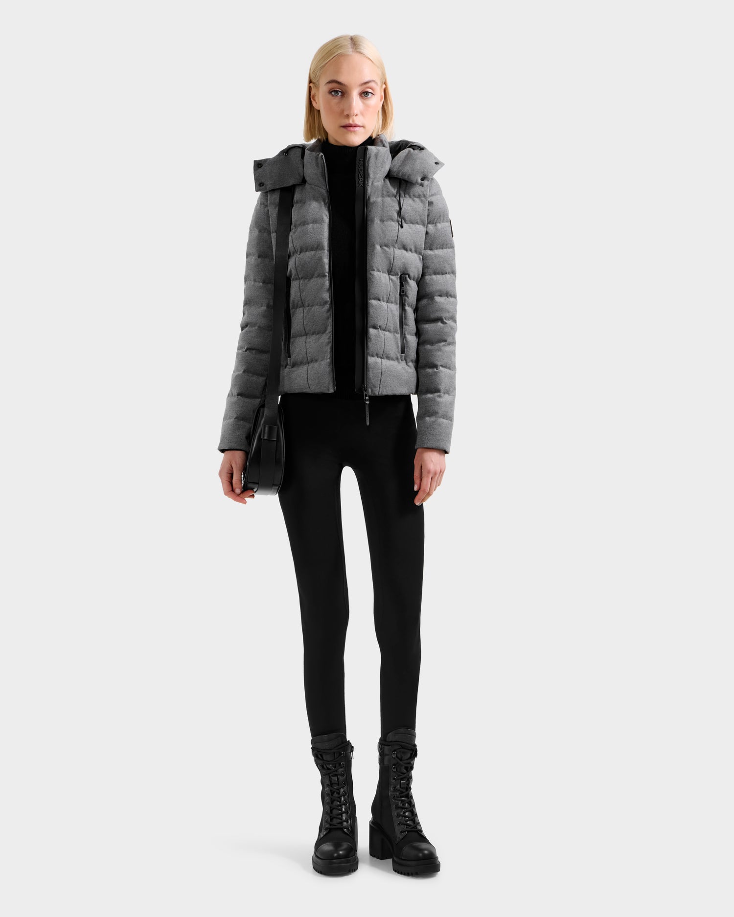 A person stands facing forward, wearing the Rudsak MELISSA W - 8125564 Heather Charcoal lightweight down jacket with black pants, black lace-up boots, and a black shoulder bag against a plain light background.