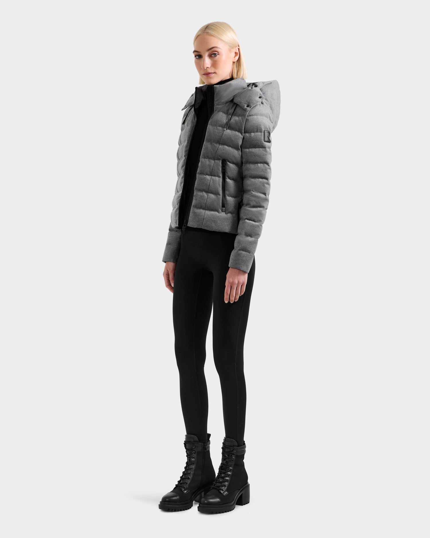 A woman with straight blonde hair and a neutral expression wears the Rudsak MELISSA W - 8125564 Heather Charcoal, a gray lightweight down jacket with a hood—perfect transitional outerwear for shifting seasons.