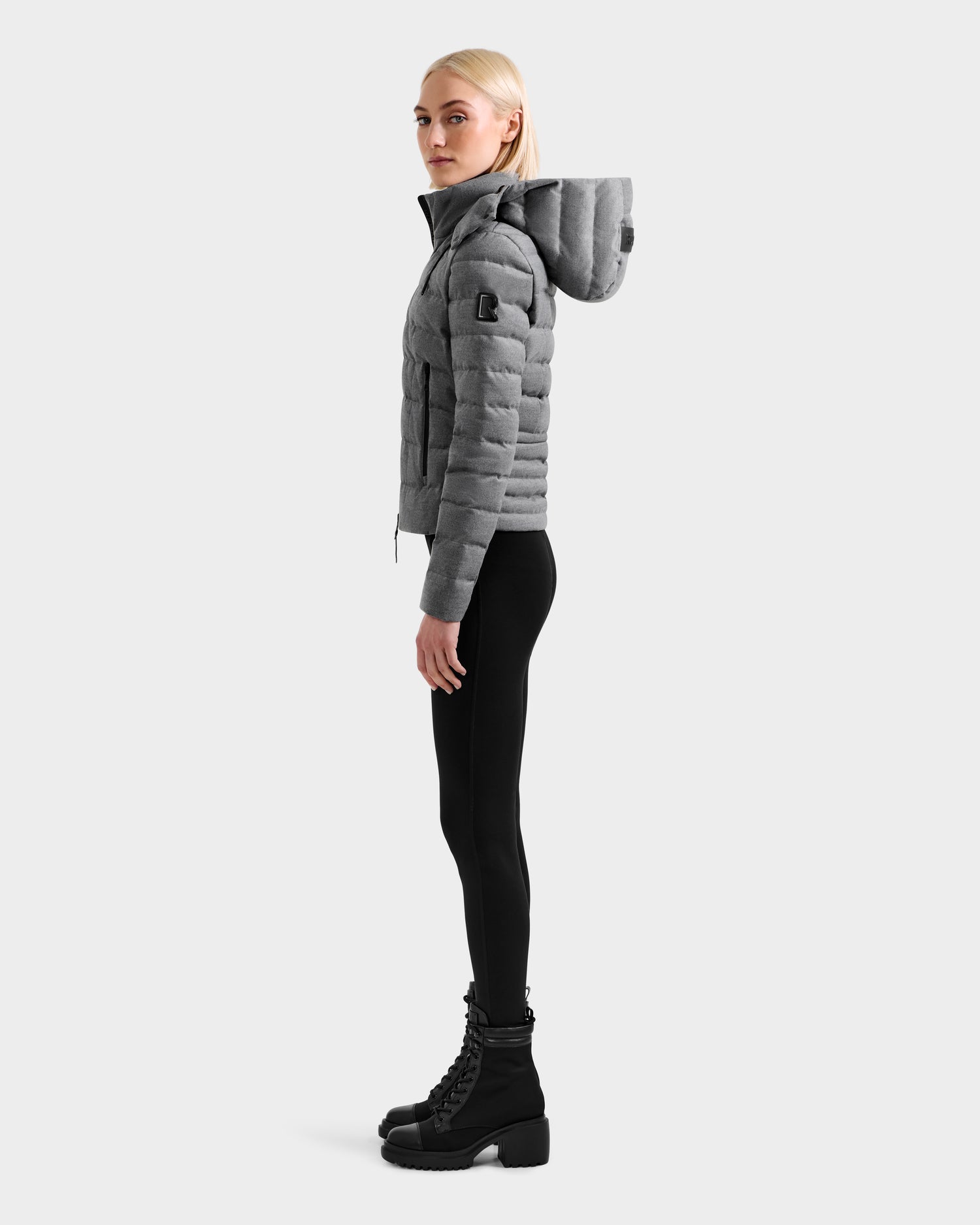 A woman stands in profile against a plain white background, wearing the Rudsak MELISSA W - 8125564 Heather Charcoal lightweight down jacket with a hood, paired with black leggings and black lace-up boots.