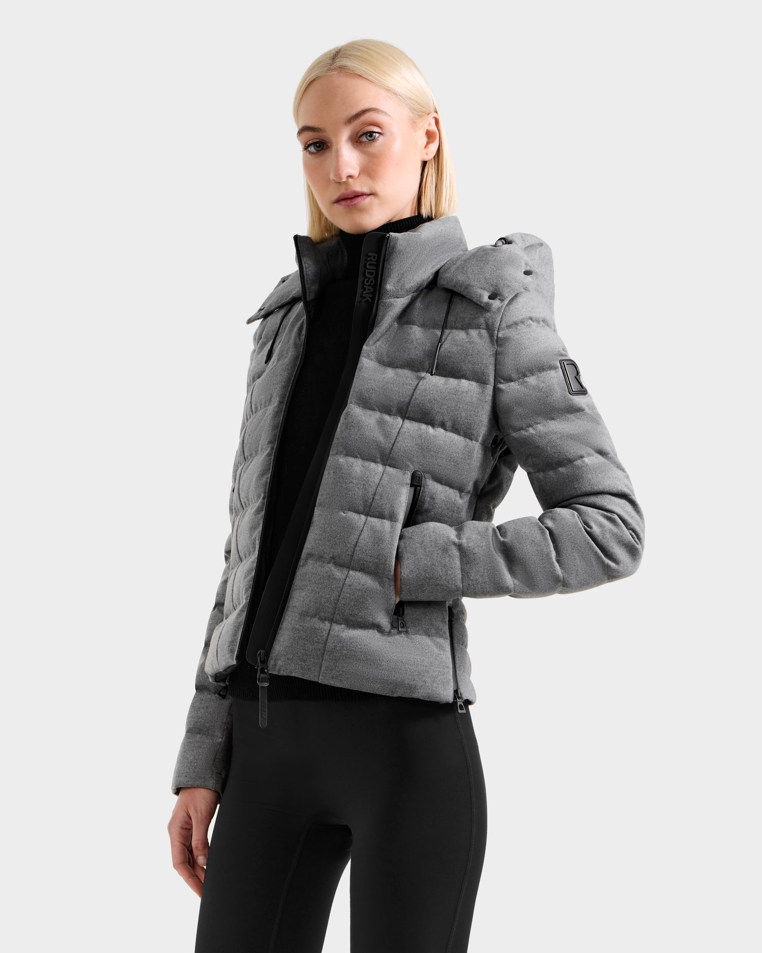 A woman with straight blonde hair wears the Rudsak MELISSA W - 8125564 Heather Charcoal lightweight down jacket over a black turtleneck and leggings, standing against a light gray background—ideal for cooler days.