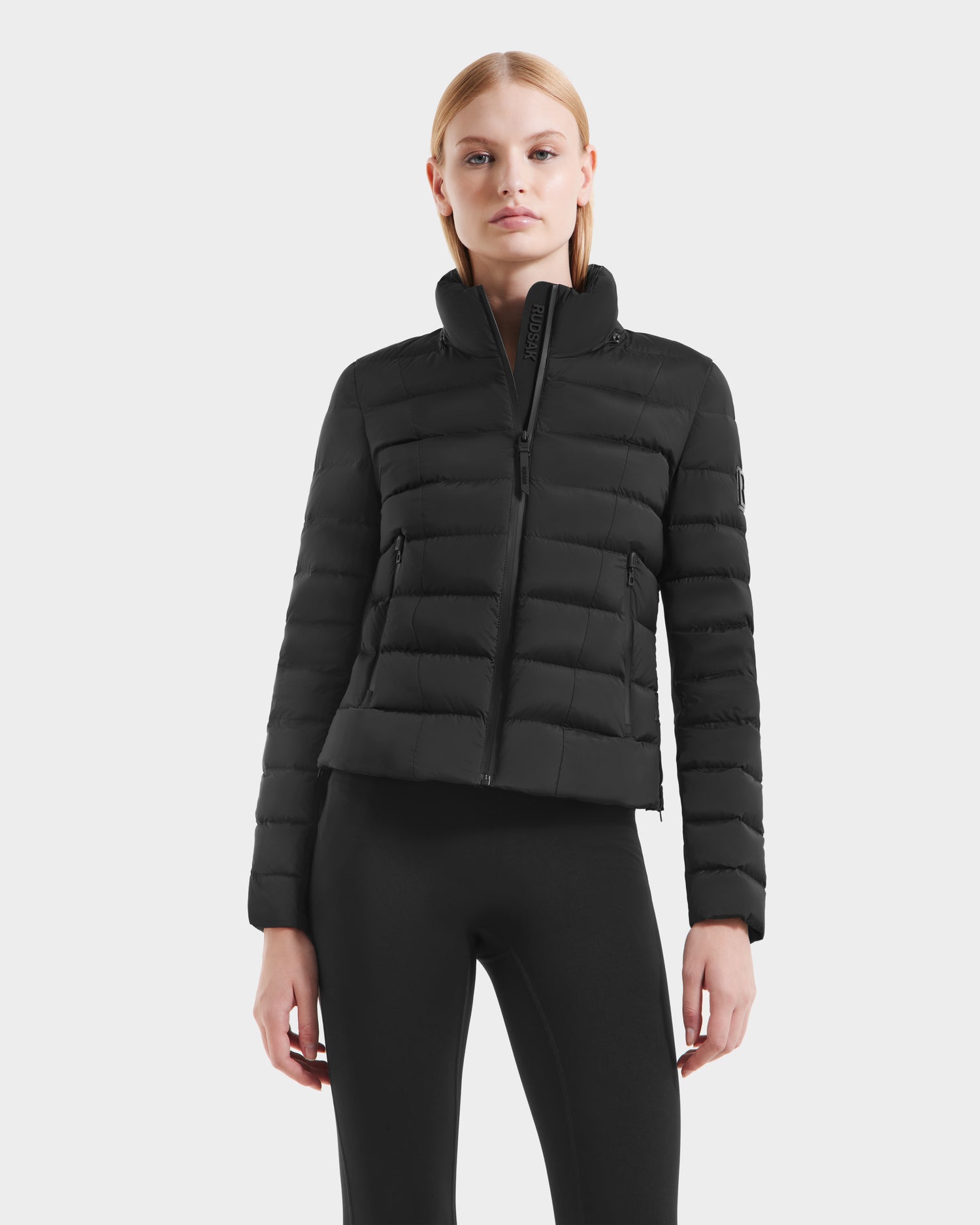 A woman with straight blonde hair wears the Rudsak MELISSA - 8125054 Black fitted womens puffer jacket and black leggings, standing against a plain white background.