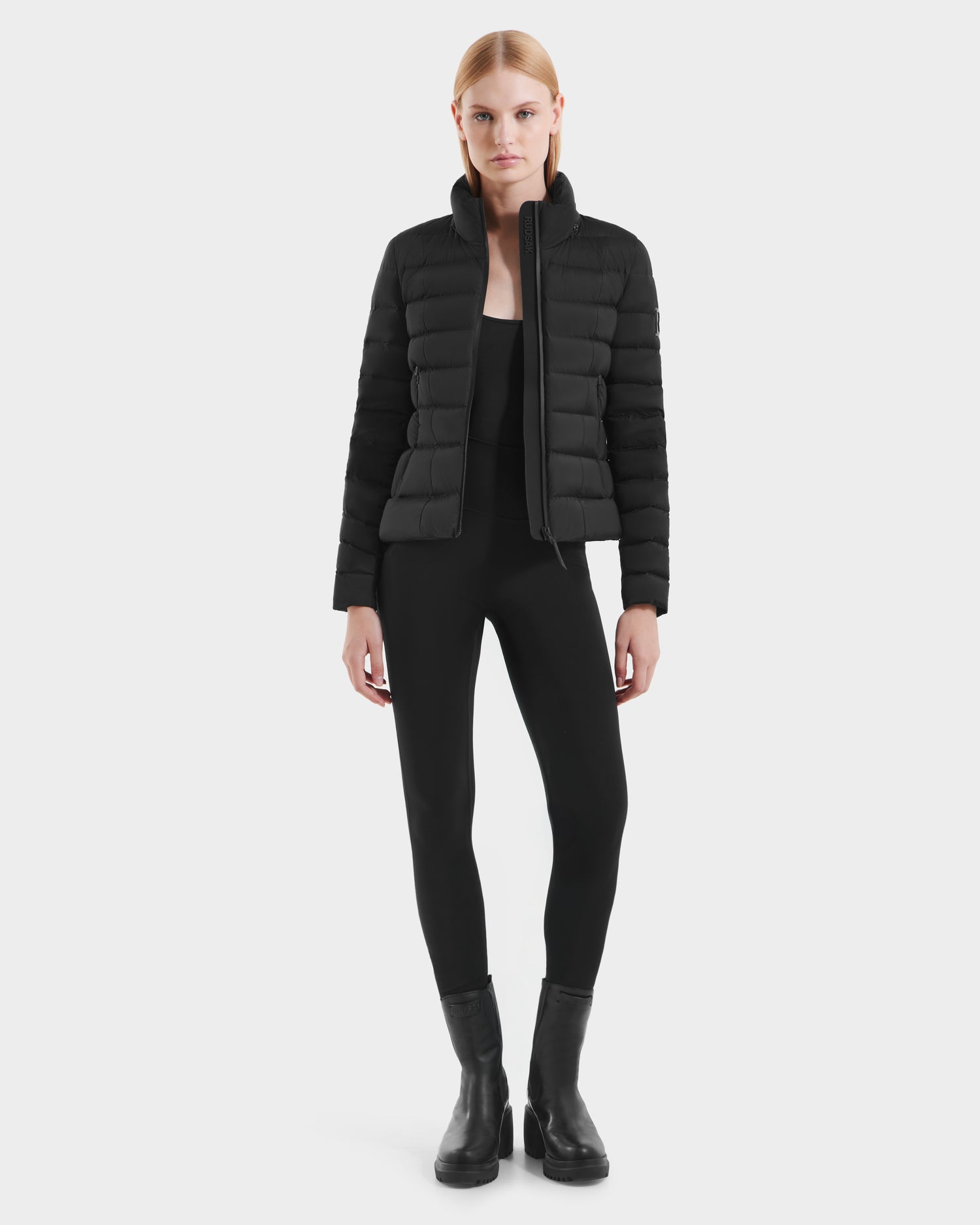 A woman stands facing forward against a plain white background, wearing the Rudsak MELISSA - 8125054 Black water-resistant women's puffer jacket, paired with black leggings and black ankle boots.