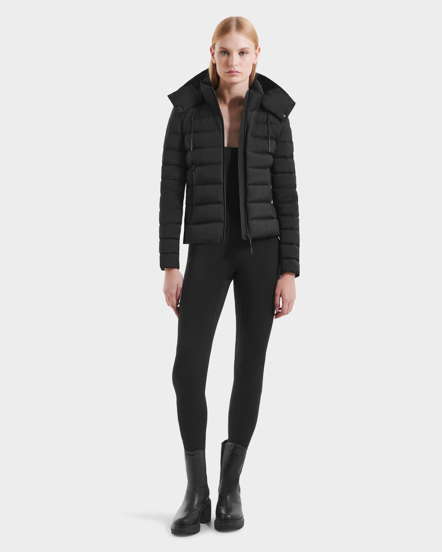 A person stands facing forward wearing the Rudsak MELISSA - 8125054 Black women's water-resistant puffer jacket with black fitted pants and black ankle boots against a plain white background.