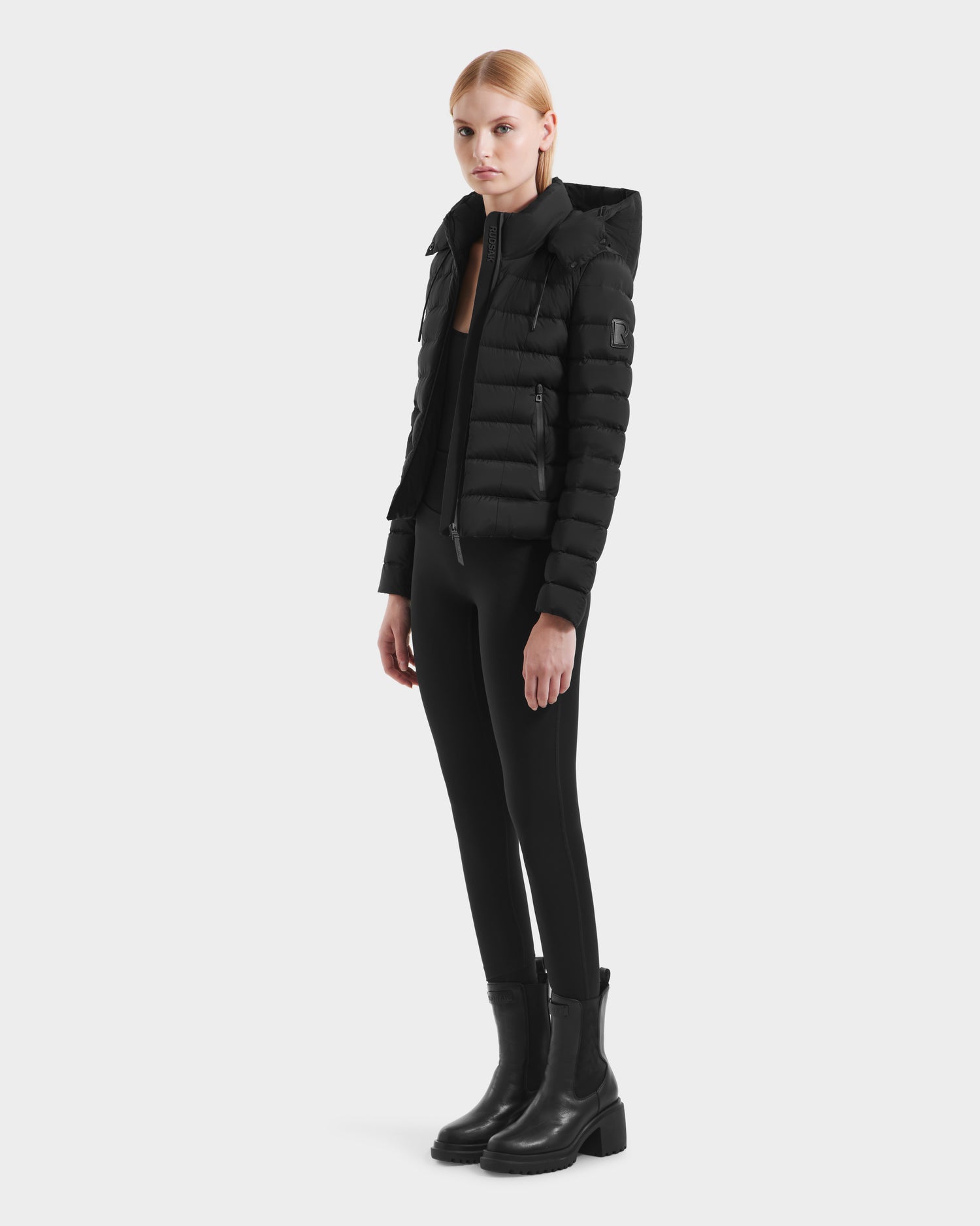 A woman with straight blonde hair pulled back wears the Rudsak MELISSA - 8125054 Black puffer jacket with RDS certified down, paired with black leggings and ankle boots, standing against a plain background with a neutral expression.