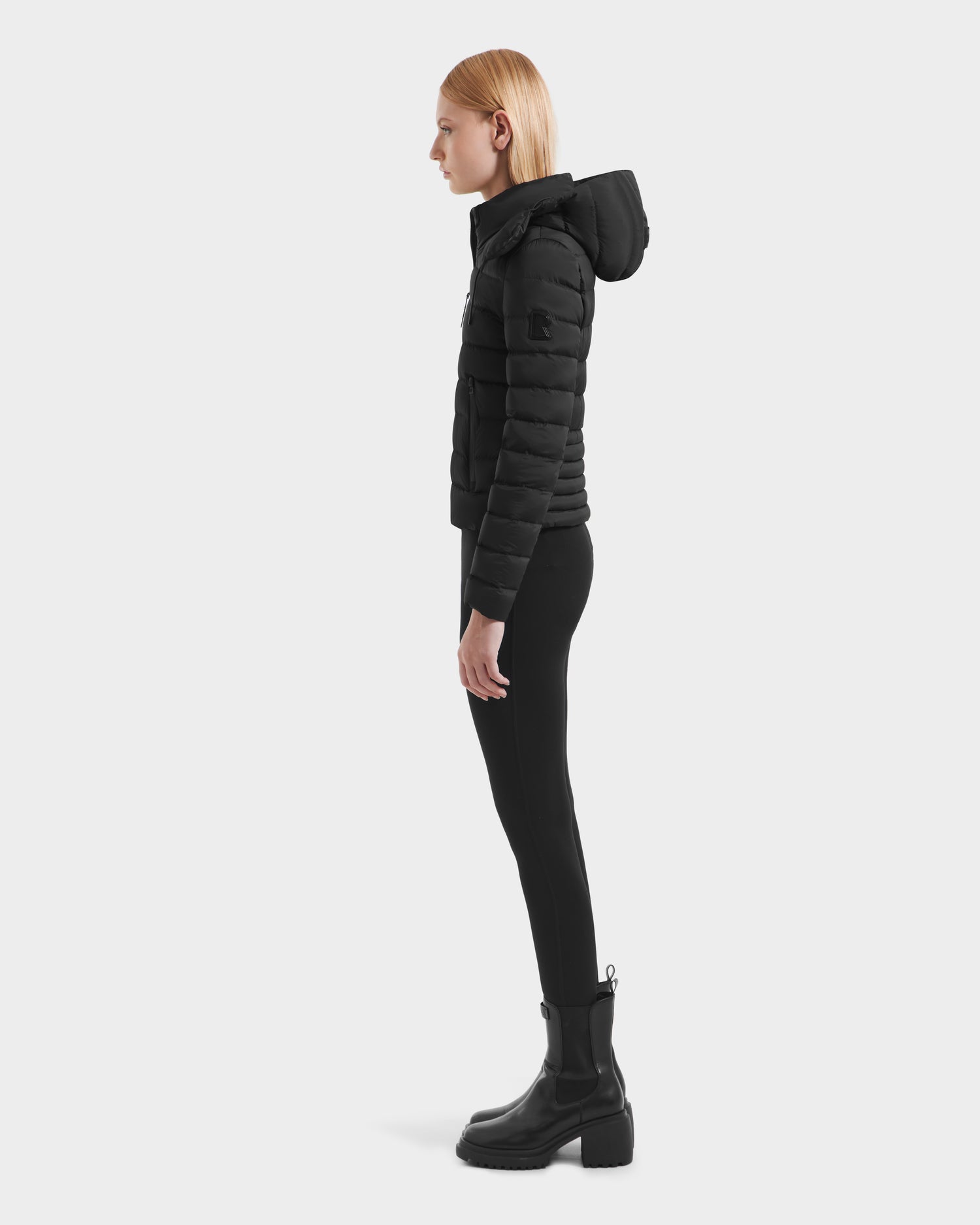 A woman stands in profile in the Rudsak MELISSA - 8125054 Black womens puffer jacket made with RDS certified down, paired with black skinny pants and chunky-heeled boots against a plain white background.