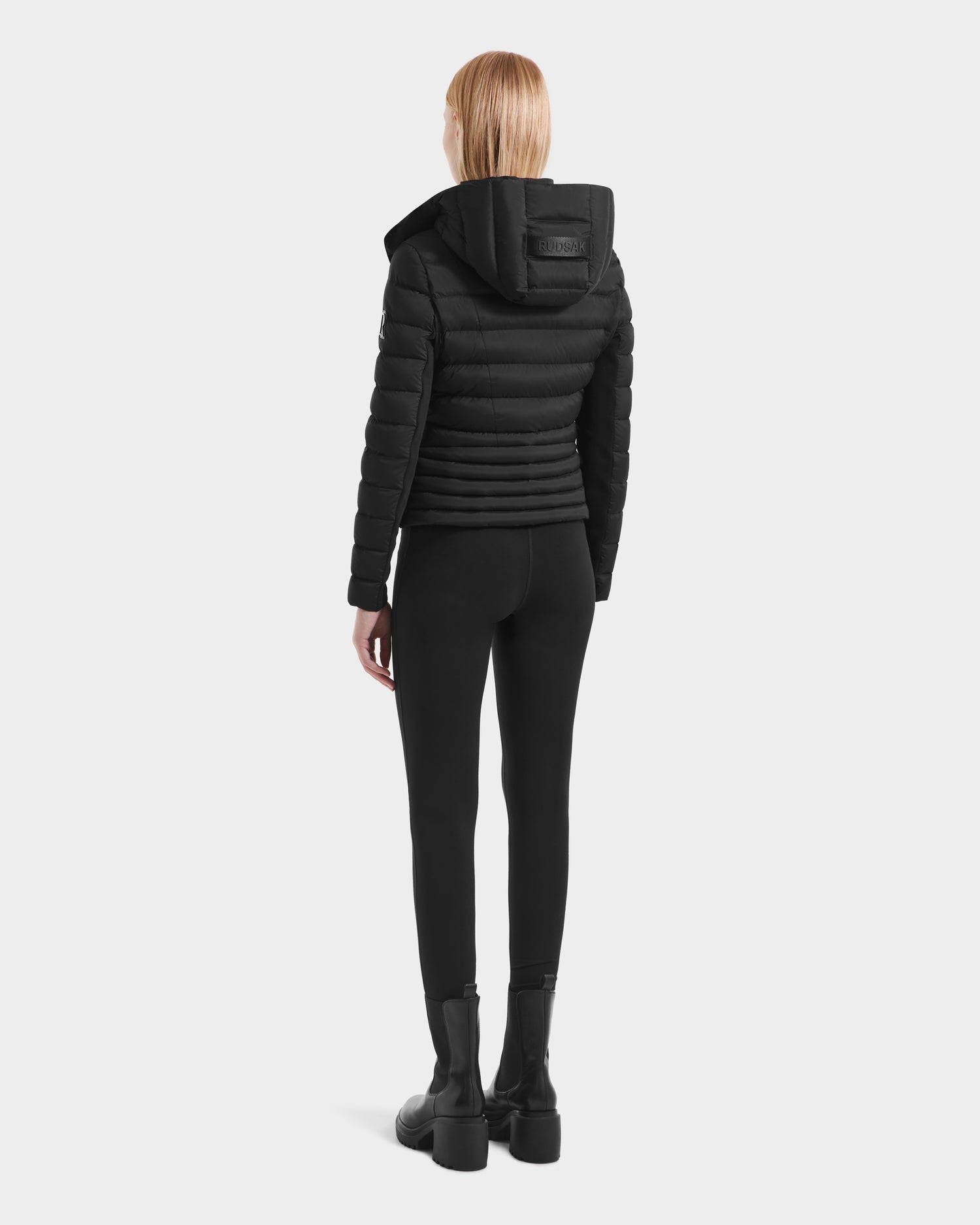A woman with straight blond hair, facing away, wears the Rudsak MELISSA - 8125054 Black womens puffer jacket, filled with RDS certified down, paired with black leggings and chunky boots against a light gray background.