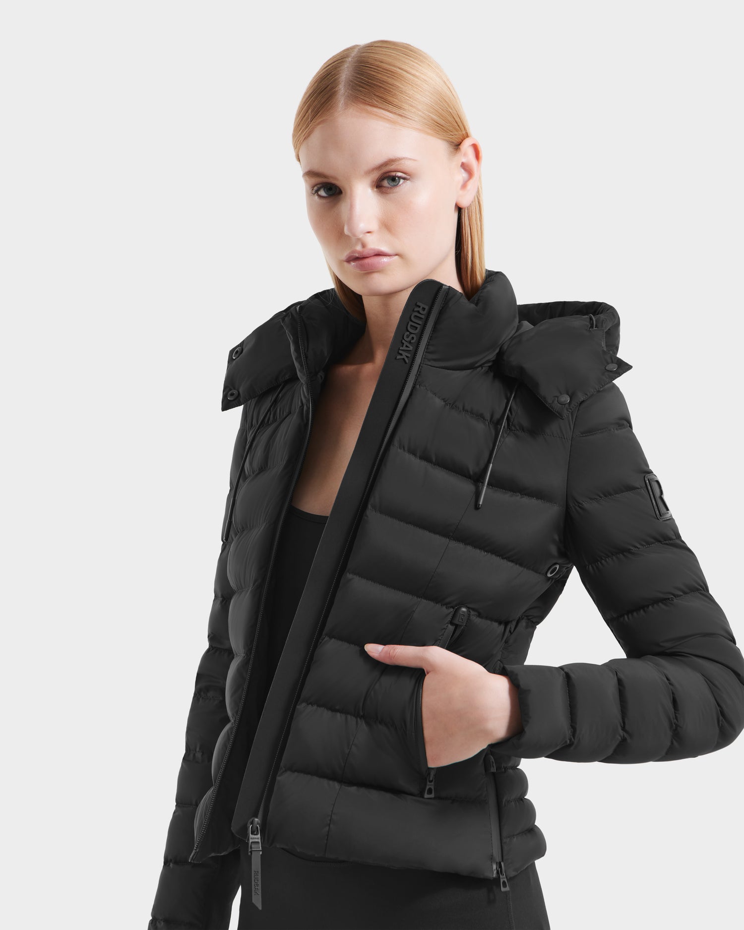 A woman with straight light blonde hair wears the Rudsak MELISSA - 8125054 Black womens puffer jacket, made from RDS certified down with a high collar and hood, partially unzipped over a black top, against a plain light gray background.